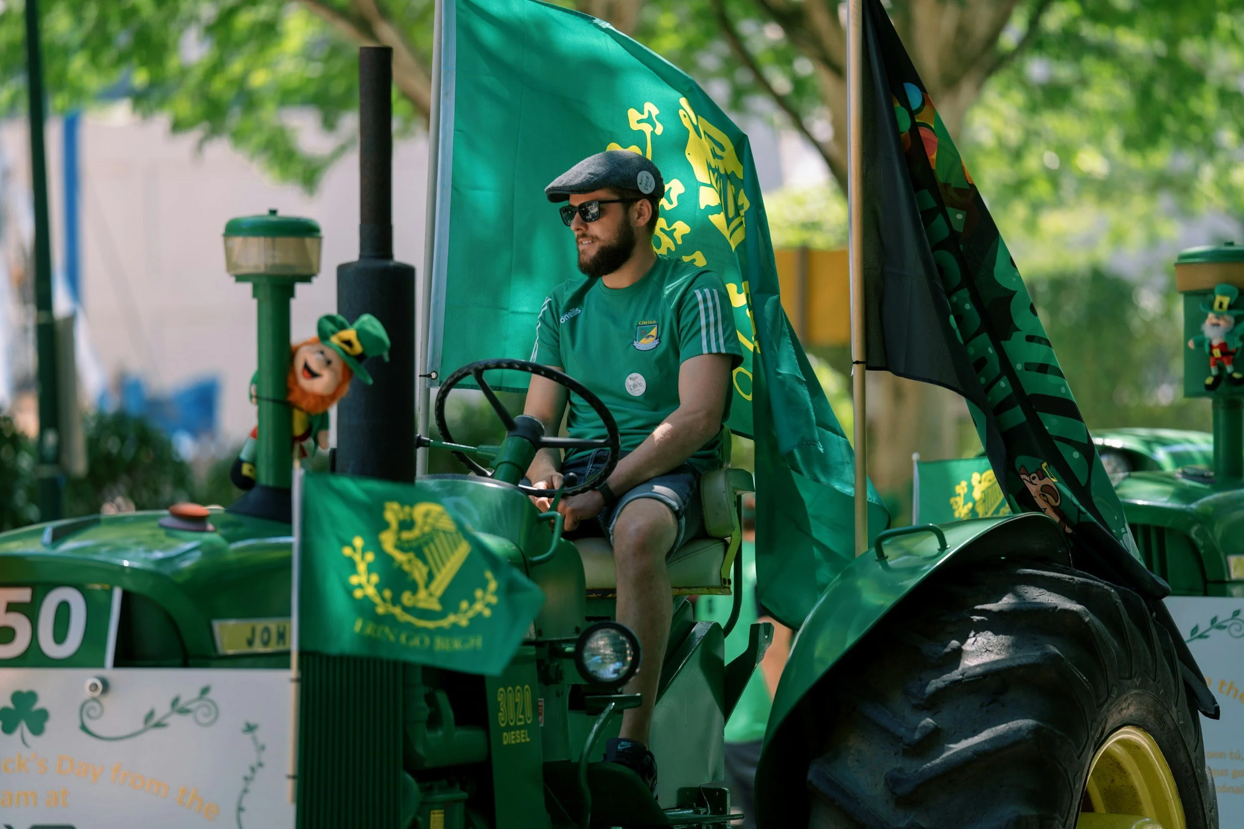 A man wearing sunglasses, a gray flat cap, and a green t-shirt with a logo, sitting on a green tractor decorated with flags and St. Patrick's Day-themed items, outdoors among trees.