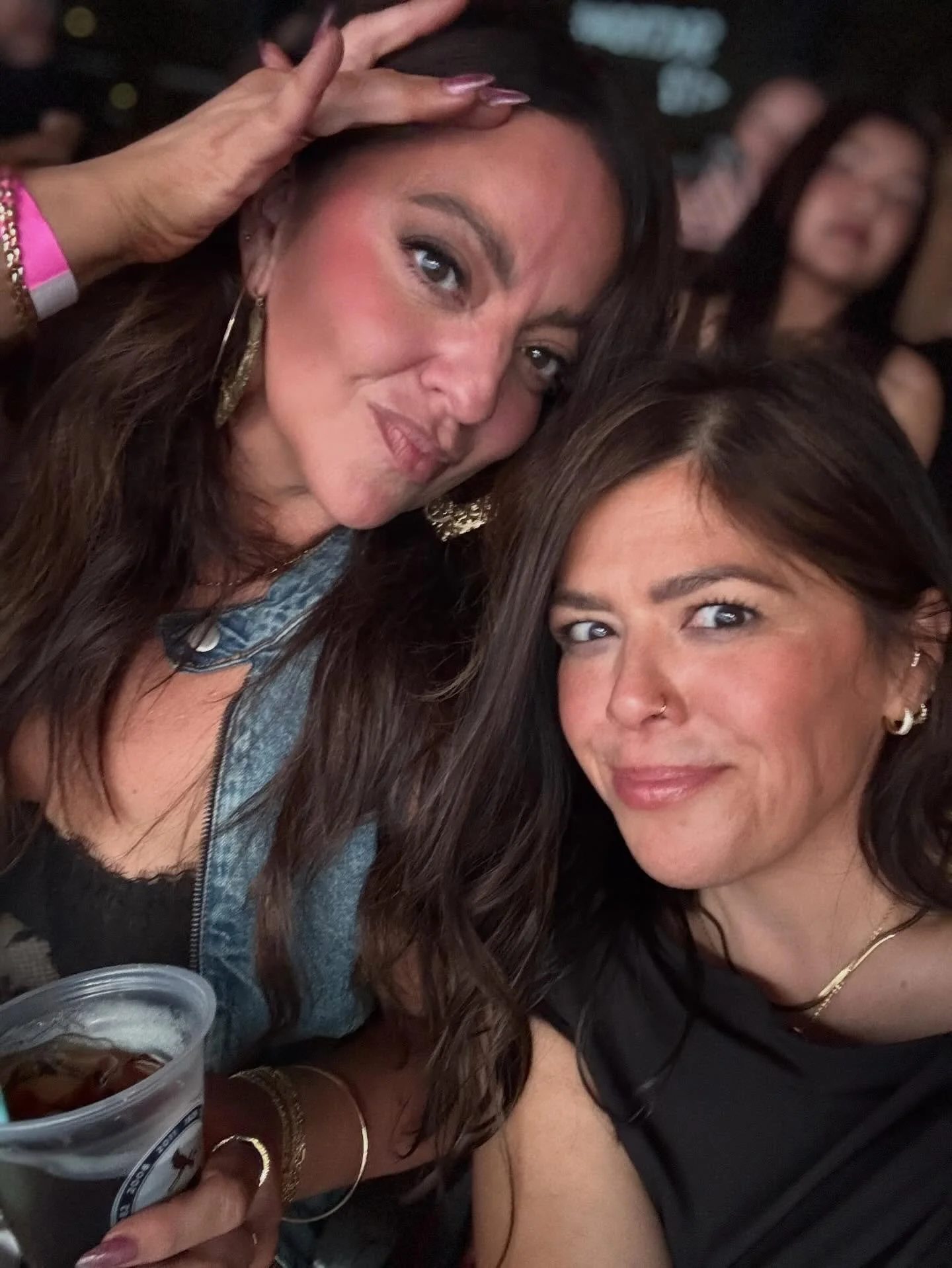 Two women taking a selfie at a social event, one woman wearing a black top and gold jewelry, the other wearing a denim jacket with a beverage in the foreground.