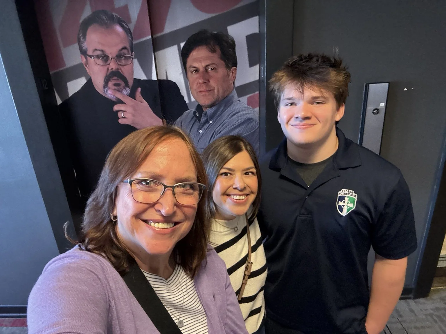 A group of three smiling people posing for a selfie in front of a poster of two men, one with glasses and a beard and the other with short dark hair, traffic signal in the background.