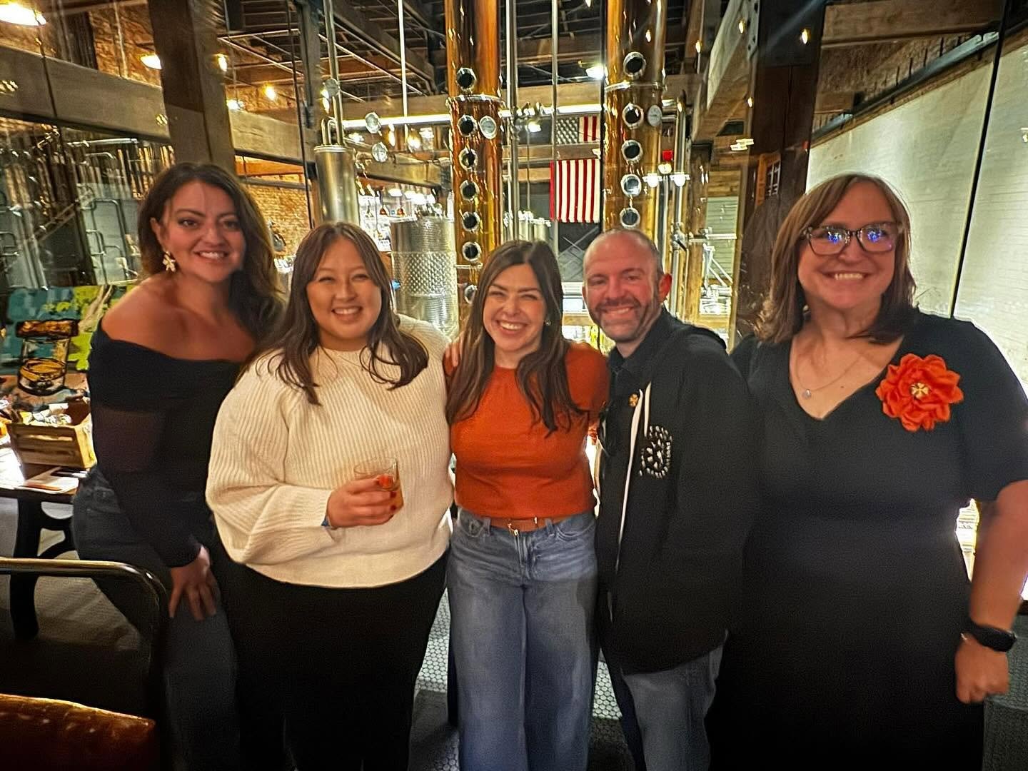 Five people smiling and standing together in a restaurant or brewery, with industrial decor including large metallic pipes and exposed brick walls in the background.
