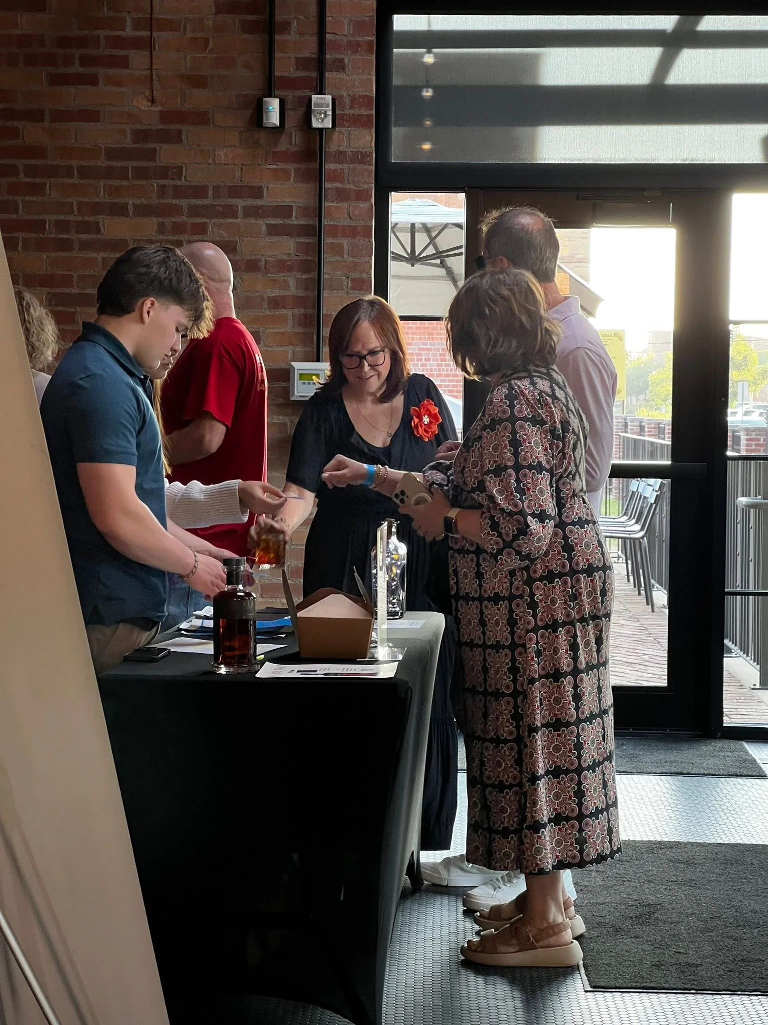 A woman in a floral dress and platform sandals is standing at a table, interacting with another woman in glasses and a black dress. Several people are gathered around, with a brick wall and large window in the background, in an indoor setting with sunlight coming in.