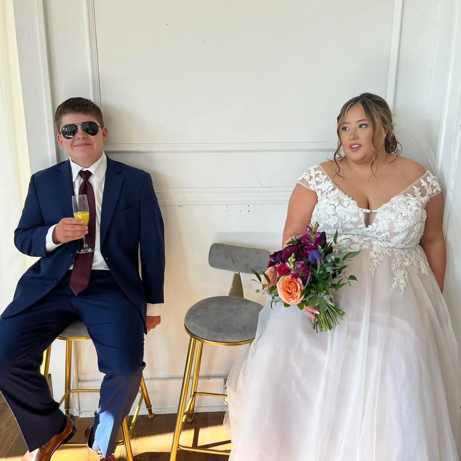 A young man in a navy blue suit and sunglasses holding a glass of champagne, sitting on a gold-stool, next to a woman in a white wedding dress holding a bouquet of flowers, both seated on chairs against a plain wall.