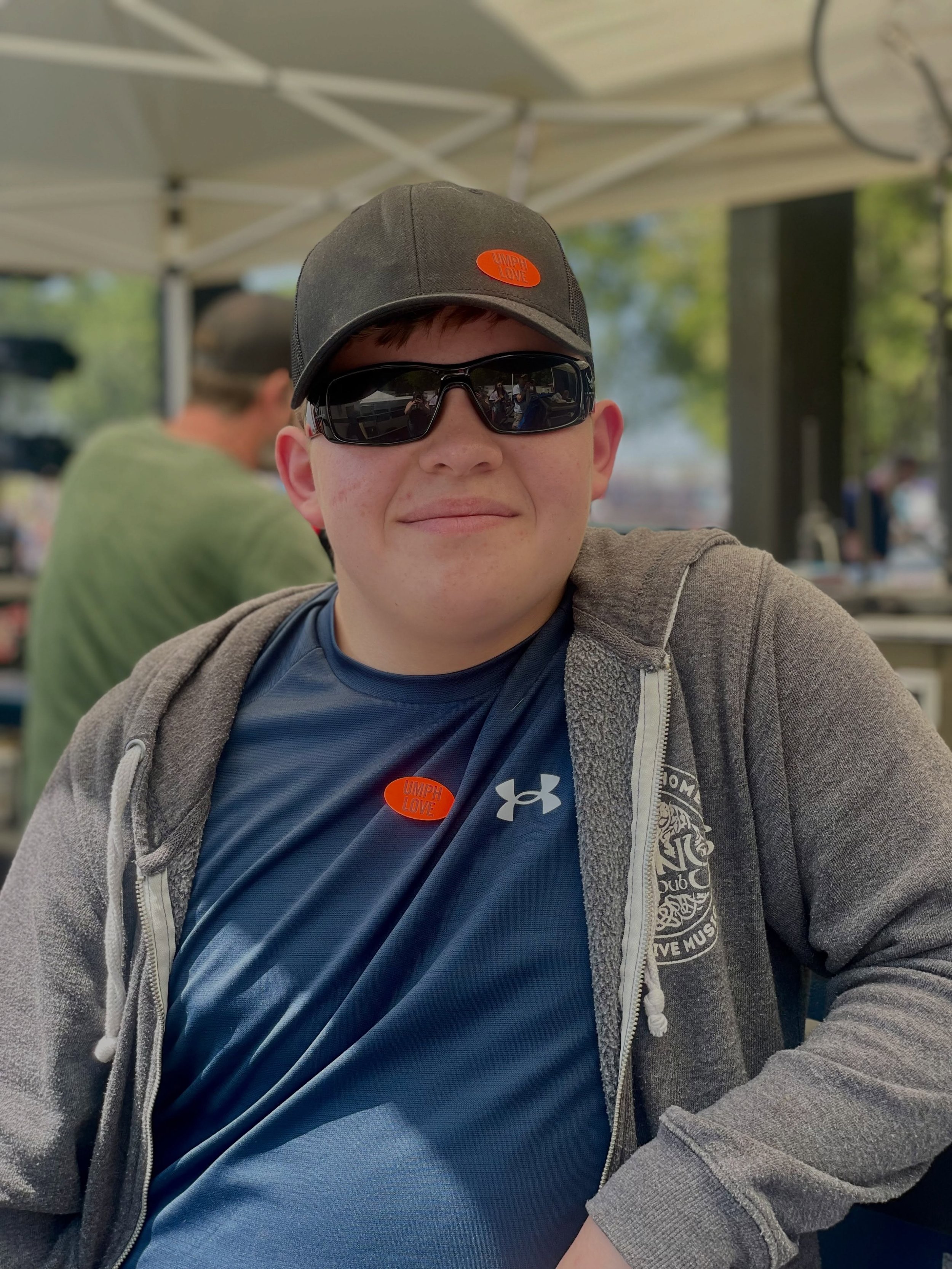 A young man wearing a gray baseball cap, black sunglasses, a blue Under Armour shirt, and a gray zip-up hoodie sitting outdoors under a white canopy.
