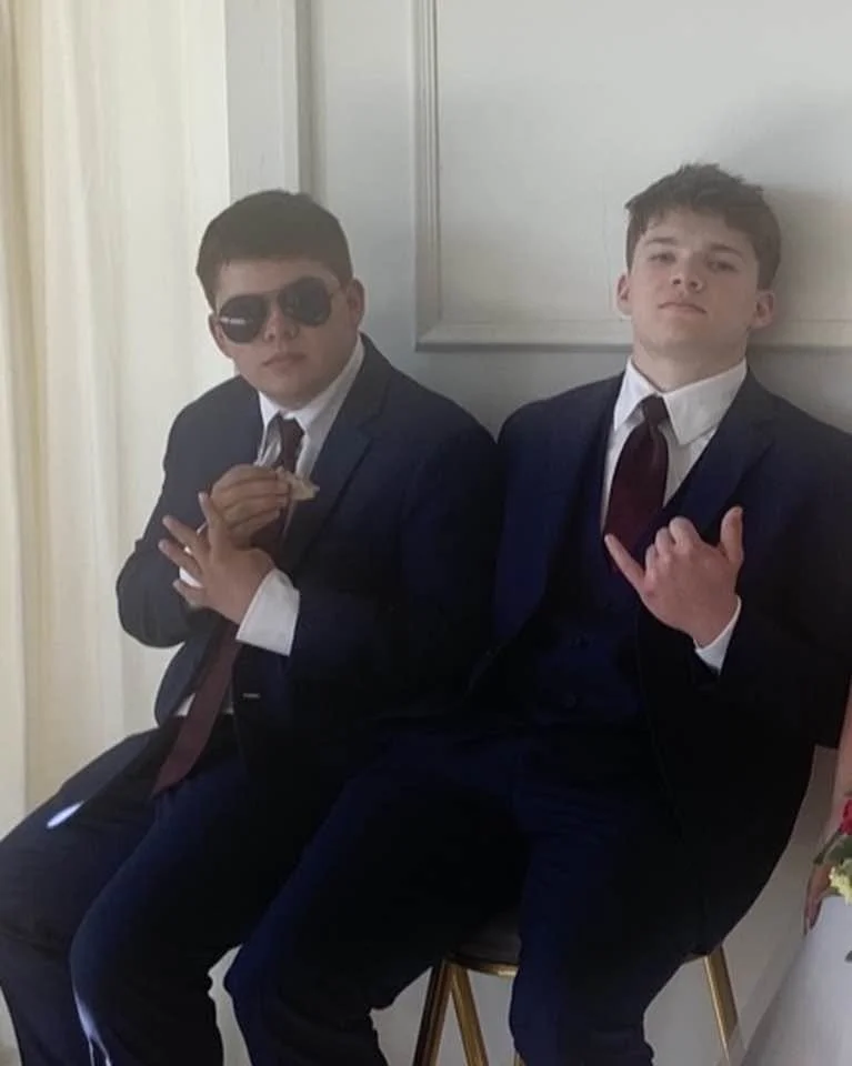 Two young men in formal suits sitting on chairs against a wall, one wearing sunglasses and holding a small snack, the other making a hand gesture.