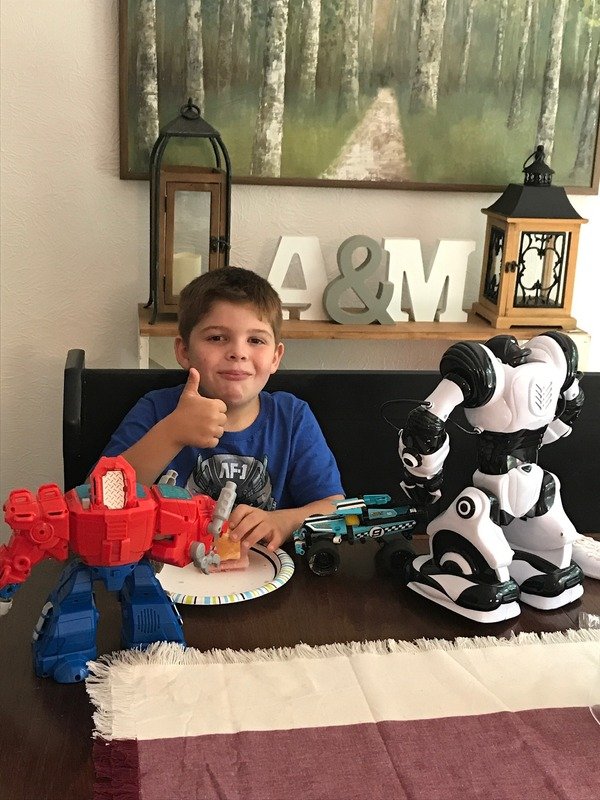 A young boy sitting at a dining table with a red and blue toy robot, two smaller cars, and a white robot dog. He is giving a thumbs-up and smiling. The background shows a wall with a picture of a forest, decorative lanterns, and large letters 'A & M' on a shelf.