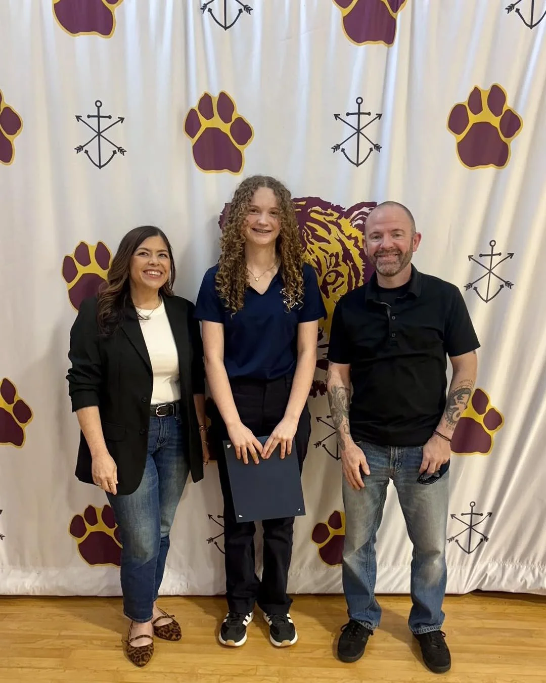 Three people standing in front of a white backdrop decorated with tiger paw prints and anchors, with a large tiger face graphic in the center.