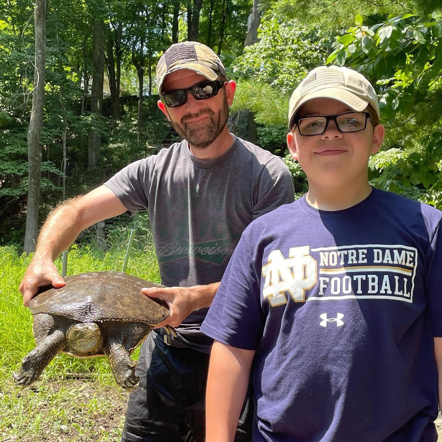 Two males outdoors in a forested area with green foliage, one holding a large turtle with a broad shell and four limbs, and the other wearing glasses and a Notre Dame football T-shirt, both smiling.