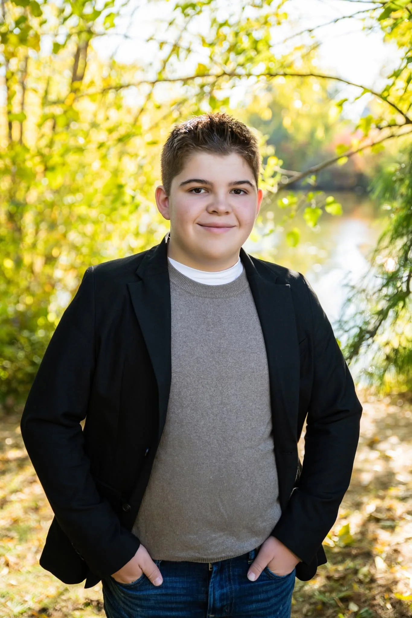 A young man with short dark hair, smiling, wearing a black jacket over a gray sweater and dark jeans, standing outdoors with fall foliage and a creek in the background.