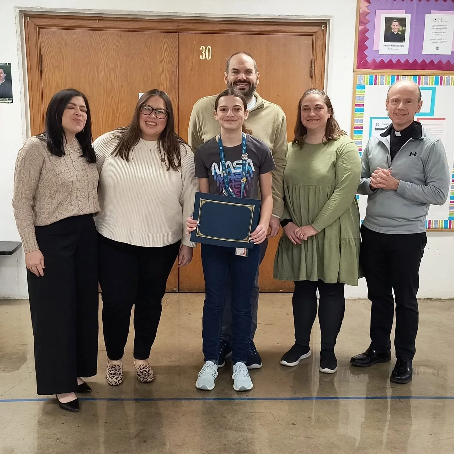 A group of six people, including a girl with a NASA T-shirt holding a certificate, standing in a school hallway in front of a door and colorful bulletin boards, smiling for a photo.
