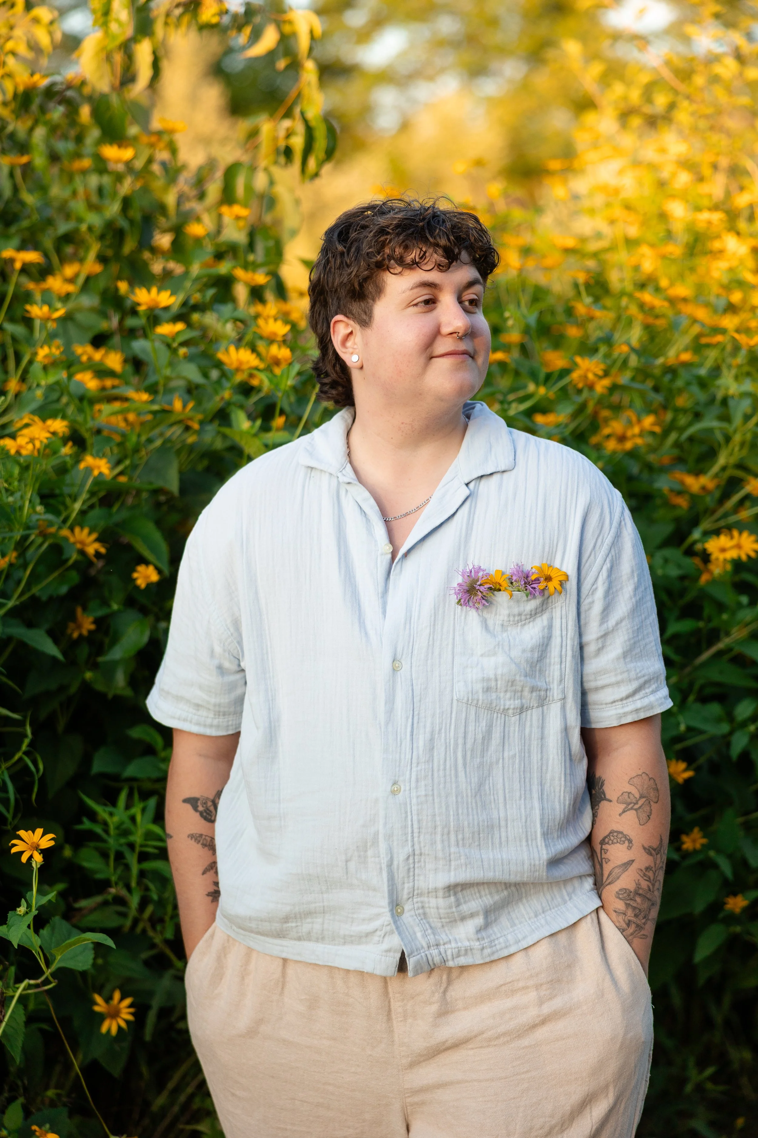 A person with short curly hair and tattoos on their arms, wearing a white button-up shirt with flowers in the chest pocket, standing outdoors in front of a backdrop of yellow flowers and greenery.
