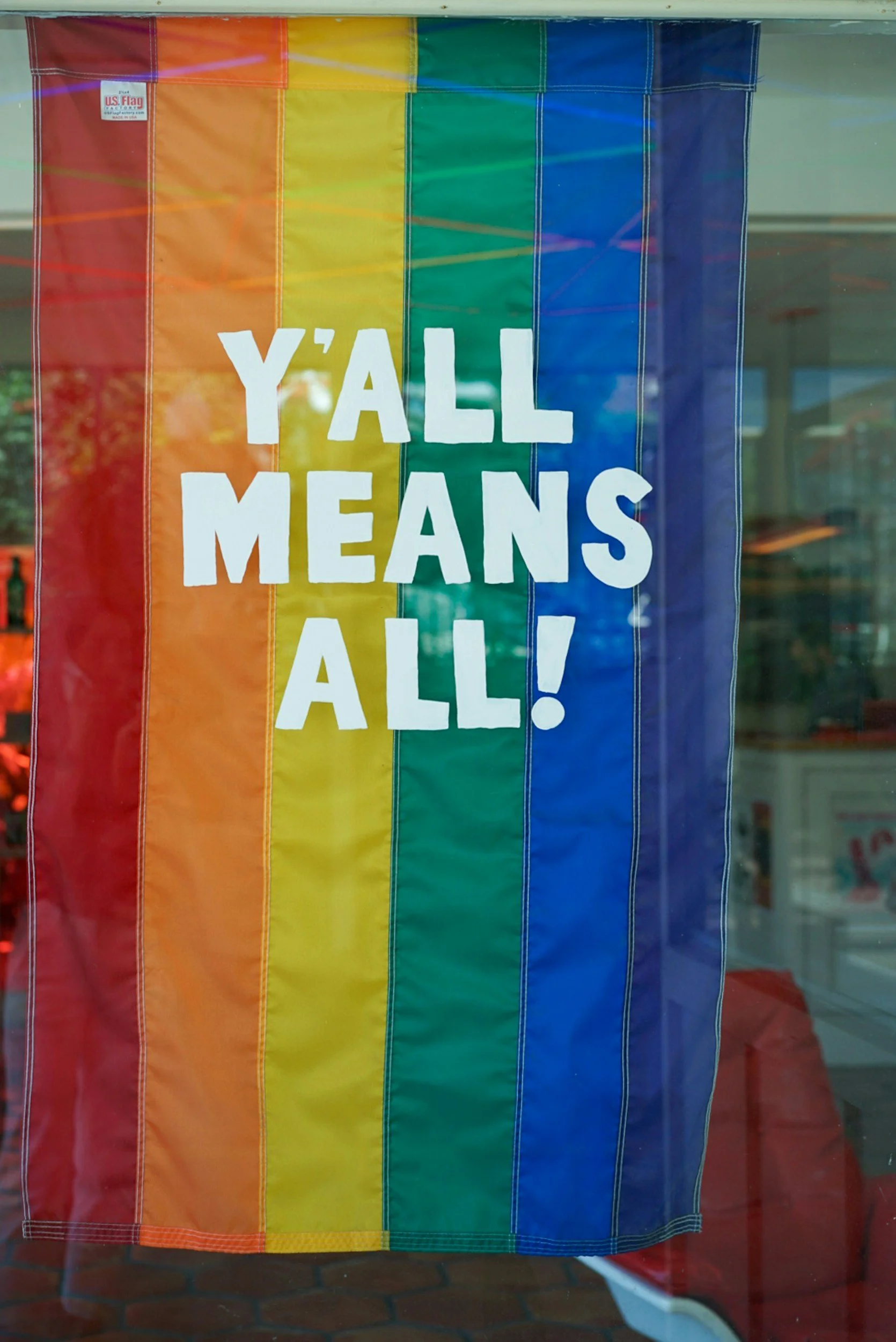 A rainbow pride flag with the phrase 'Y'ALL MEANS ALL!' written on it in white text.