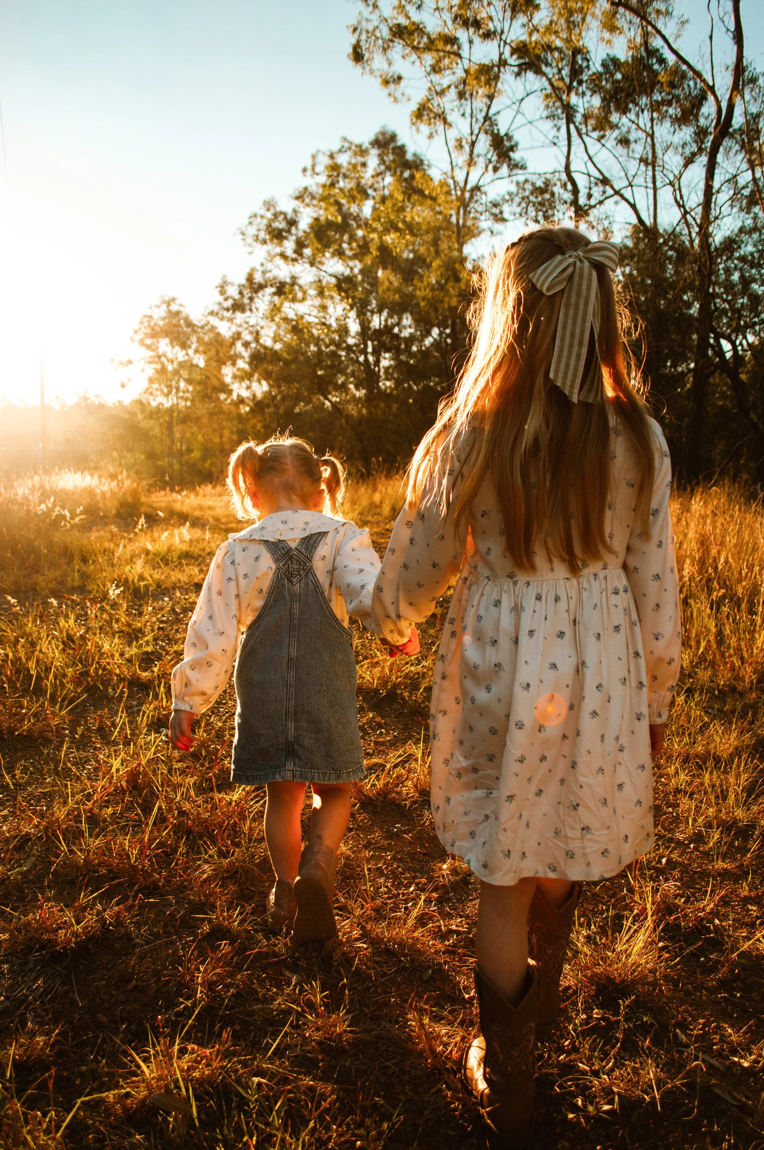 A woman and a young girl walking hand in hand through a grassy field at sunset. The woman's face is not visible, and she has long hair with a bow. The girl has pigtails and is wearing denim overalls and a long sleeve shirt. There are trees in the bac