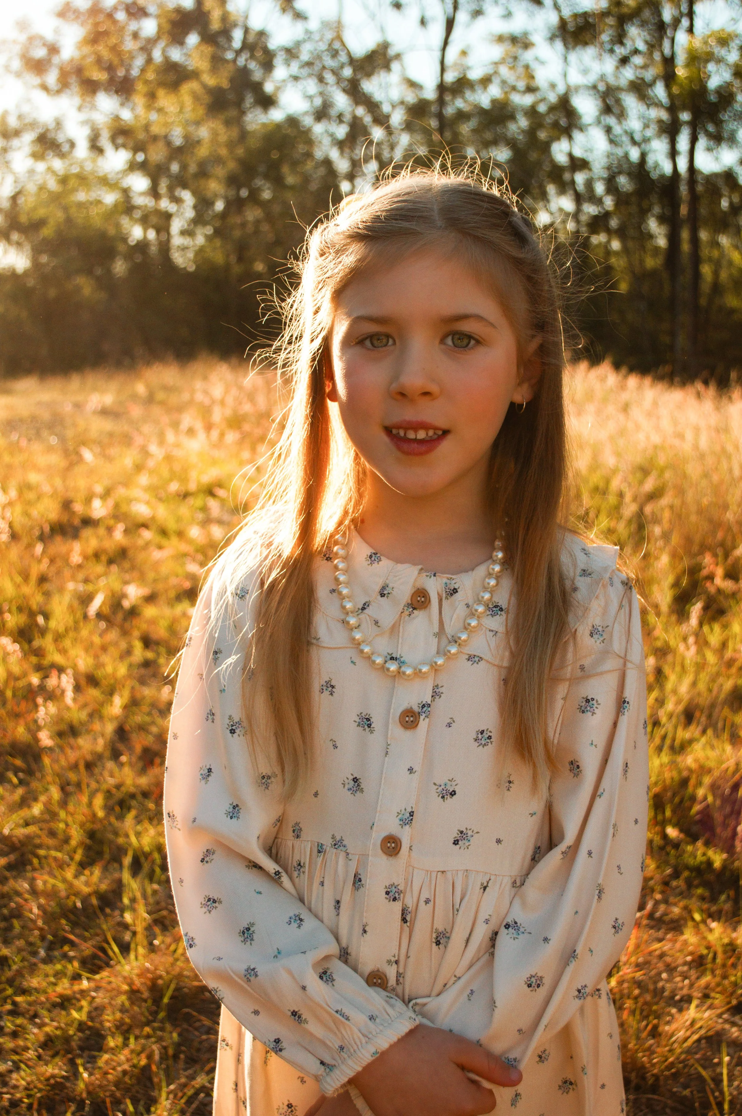 A young girl with light brown hair and hazel eyes standing in a field of golden grass during sunset. She is wearing a cream dress with small floral patterns, a pearl necklace, and has her hands clasped in front of her. Trees are visible in the backgr