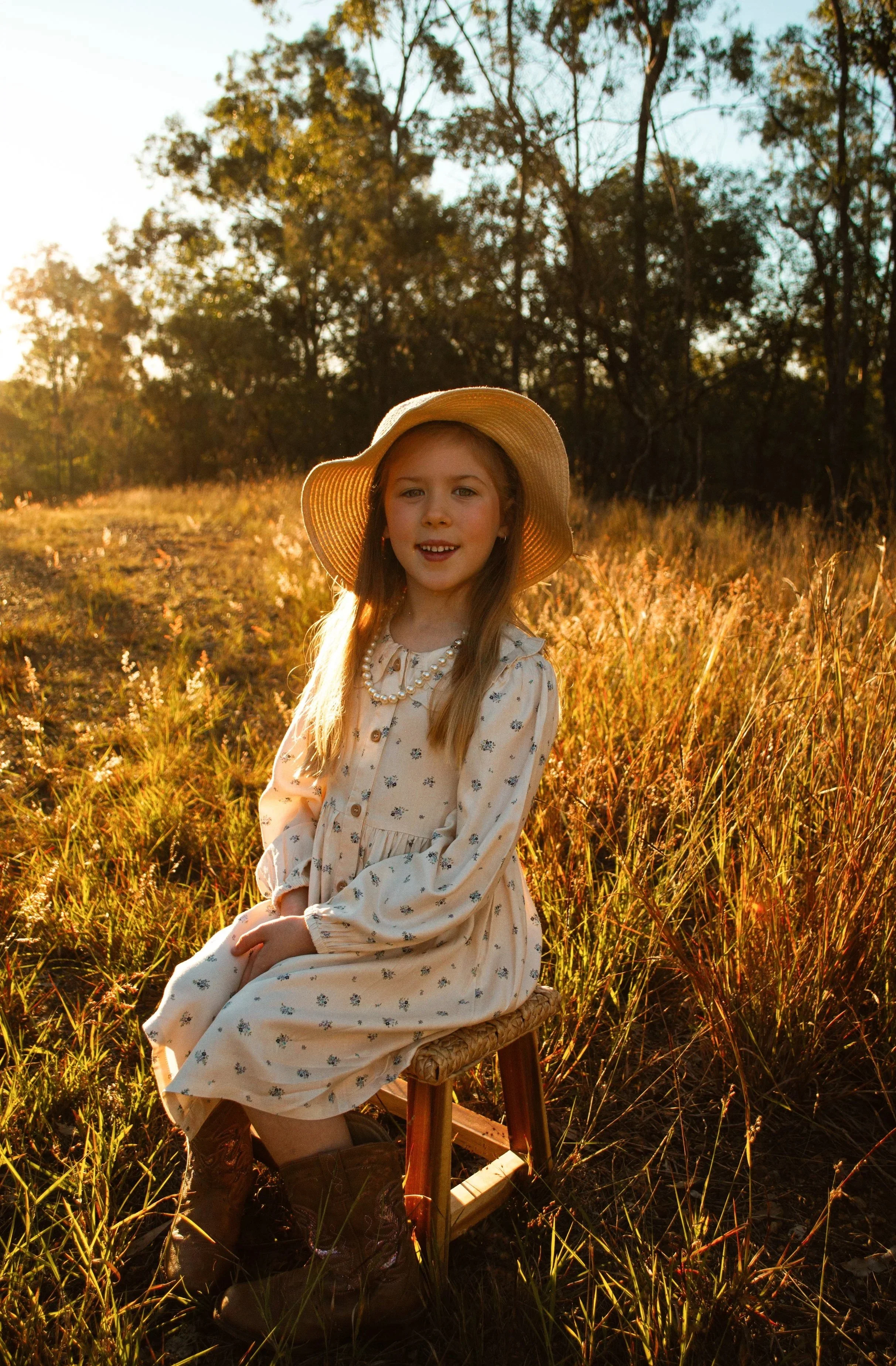 A young girl sitting outdoors in a field during golden hour, wearing a white dress with small floral patterns, a pearl necklace, and a wide-brimmed straw hat. She is smiling at the camera, with trees and sunlight in the background.
