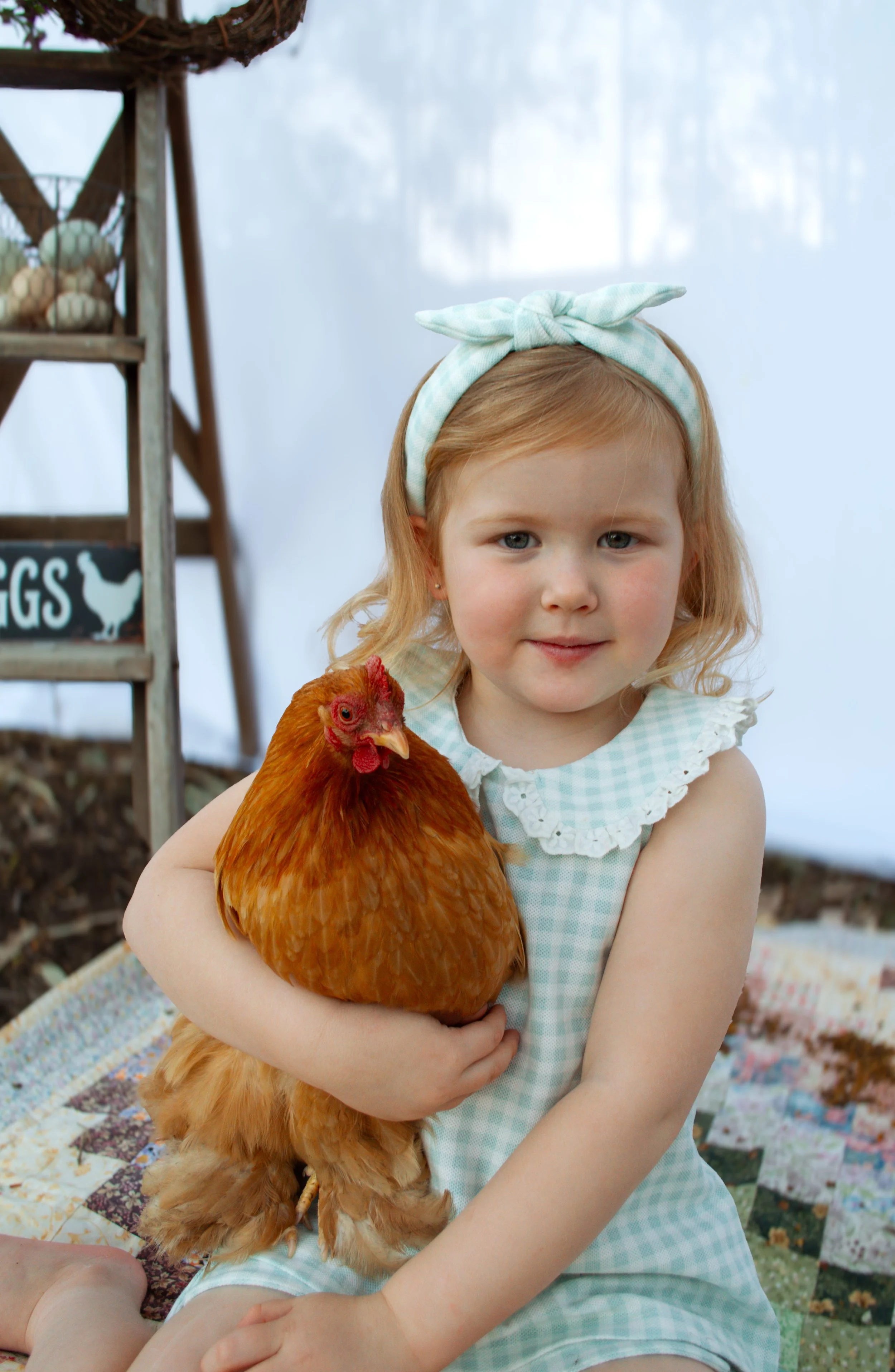 A young girl with blonde hair wearing a light blue headband with a bow, holding a large brown chicken in her arms inside a wooden henhouse with nesting boxes and a sign that says "EGGS."