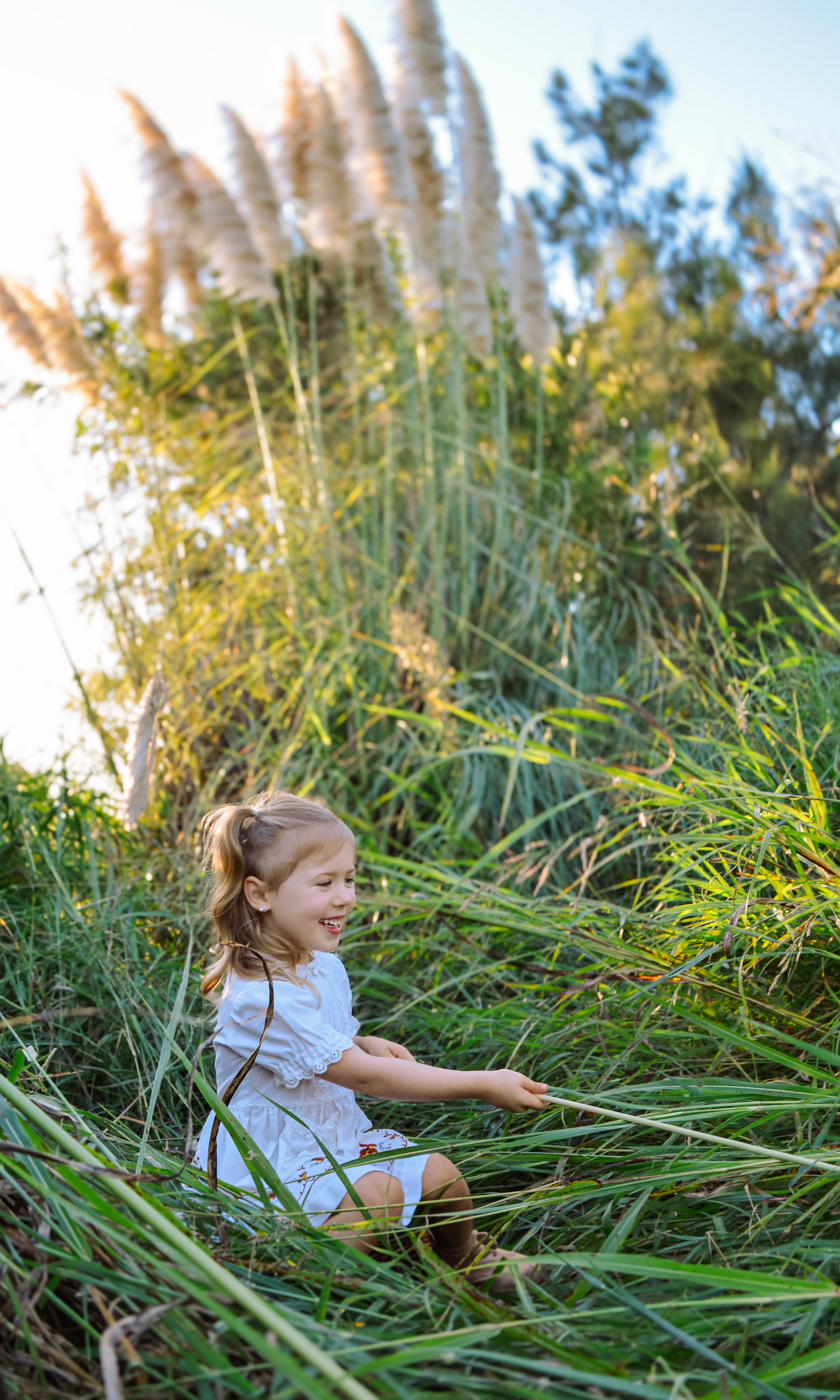 A young girl with blonde hair and a white dress sitting among tall green grass, smiling and holding a long thin stick, with large tall plants and trees in the background under a clear sky.