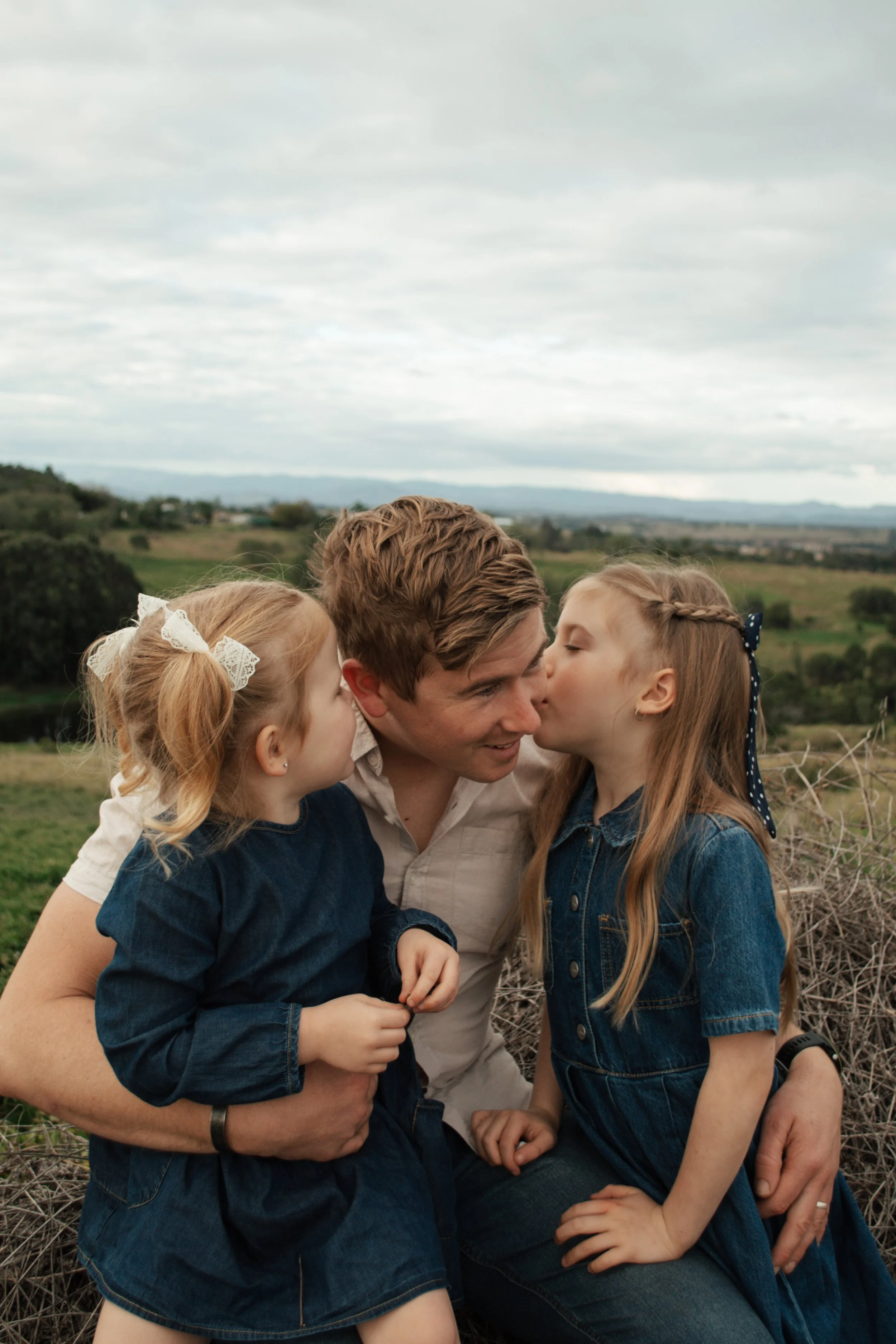 A man and two young girls are outdoors, appearing to share a moment with their faces close together and eyes closed. The background features a scenic landscape with rolling hills and a cloudy sky.