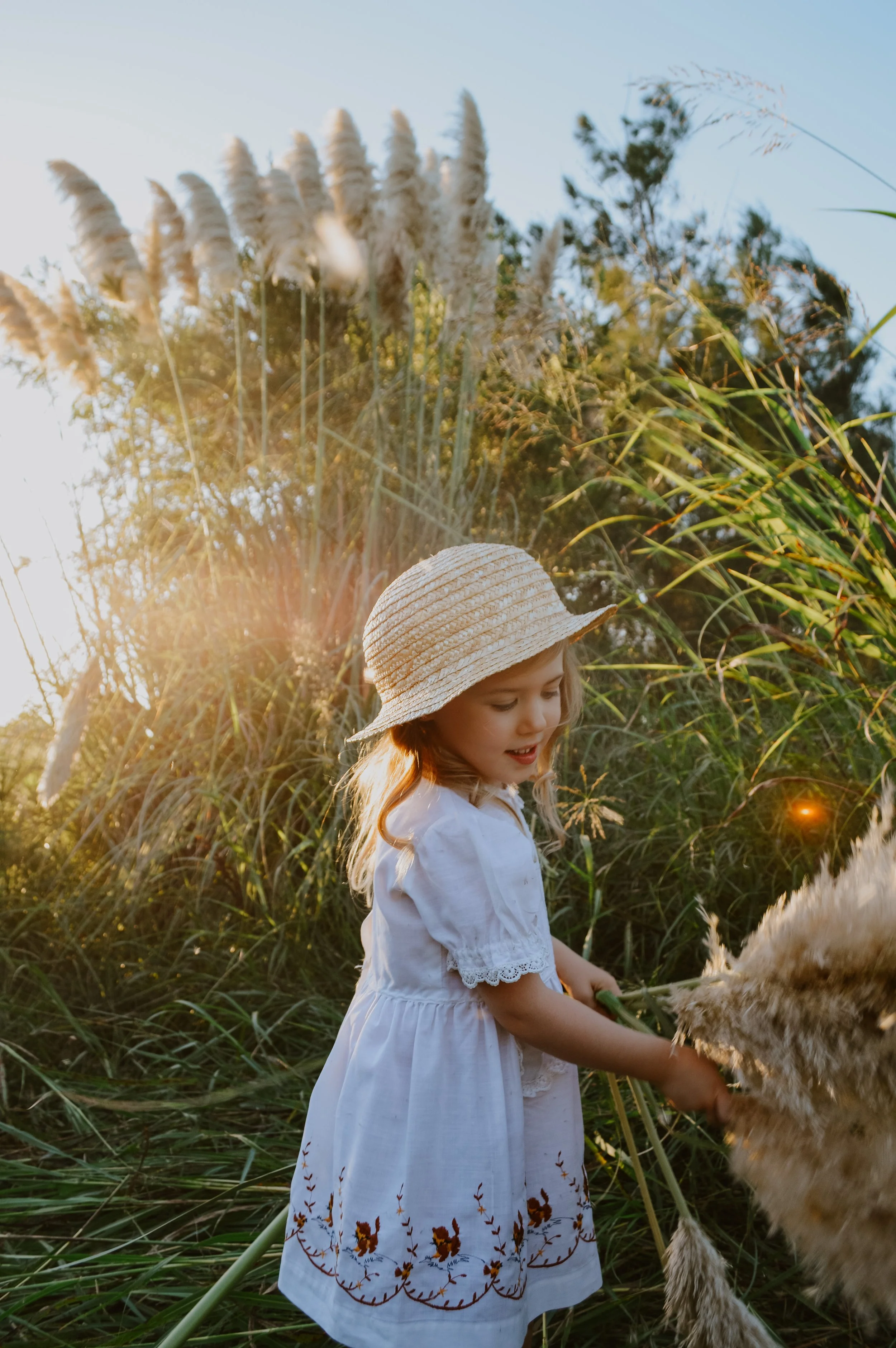 Young girl in a white dress and straw hat holding a fluffy animal, surrounded by tall grasses and trees in the late afternoon sunlight.