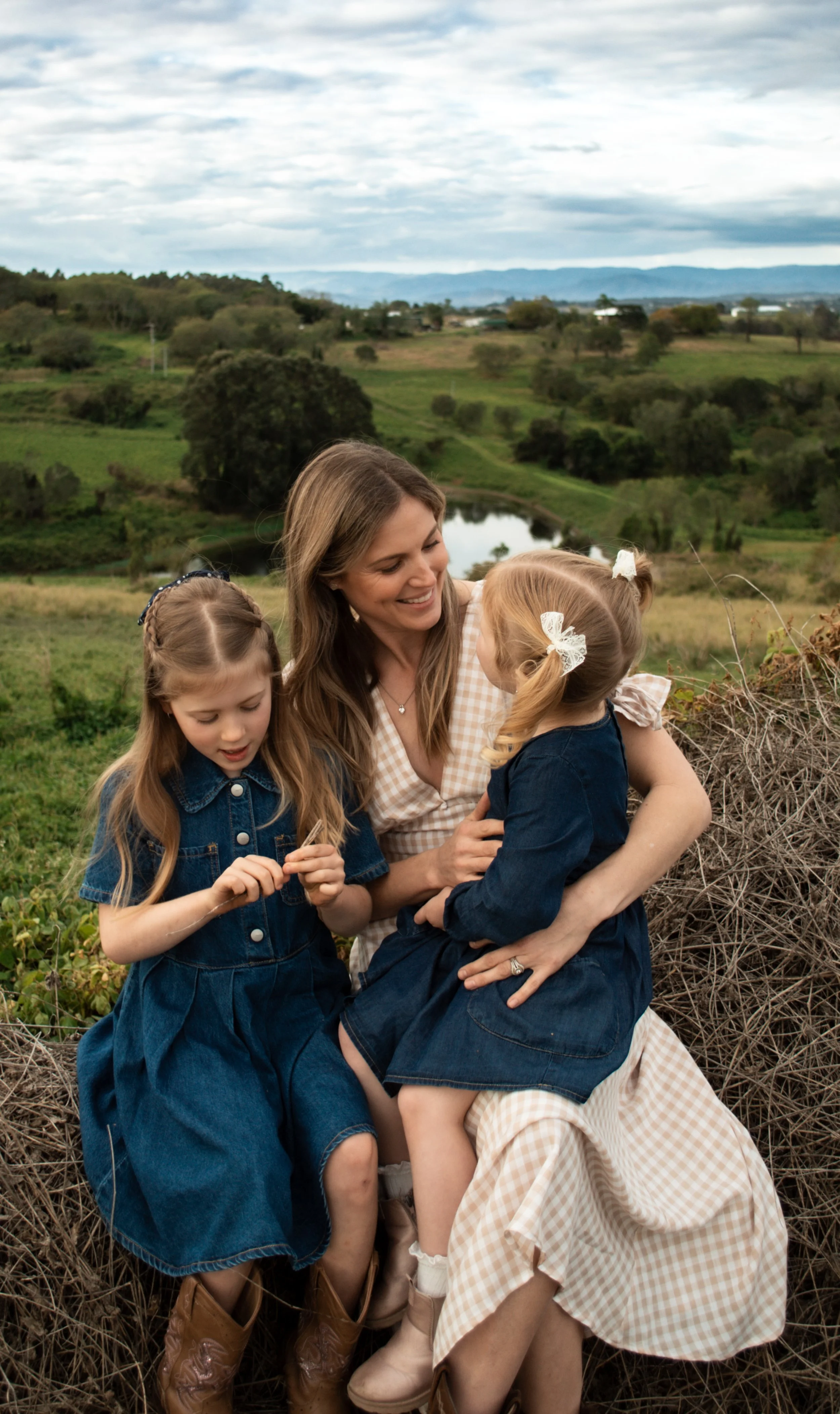 A woman and two young girls sitting on a haystack in a rural landscape with green fields, trees, and hills under a cloudy sky.