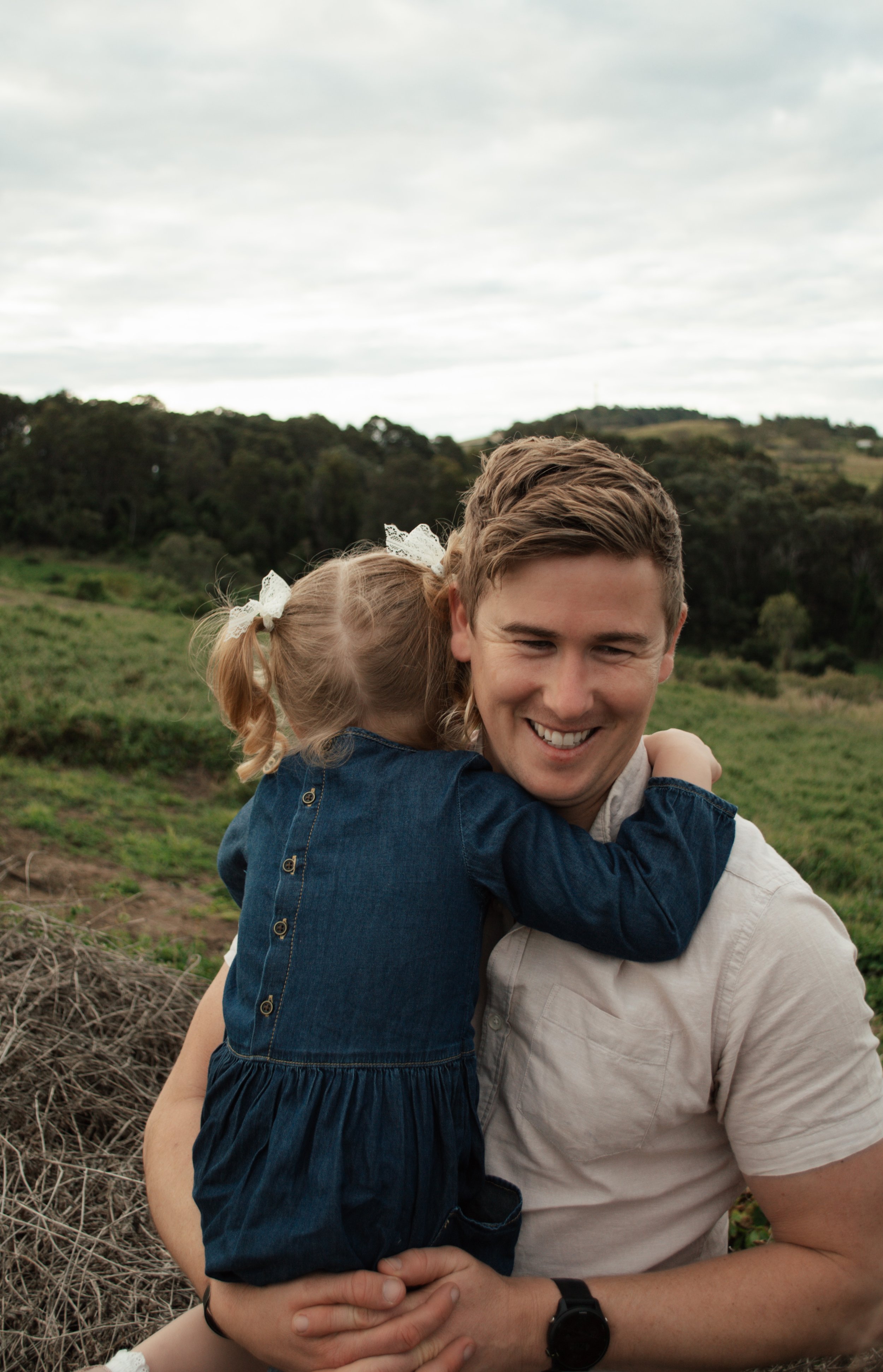 A young man with short brown hair smiling and hugging a little girl with blonde hair in pigtails, wearing a navy blue dress with buttons, outdoors in a grassy field with trees and hills under a cloudy sky.