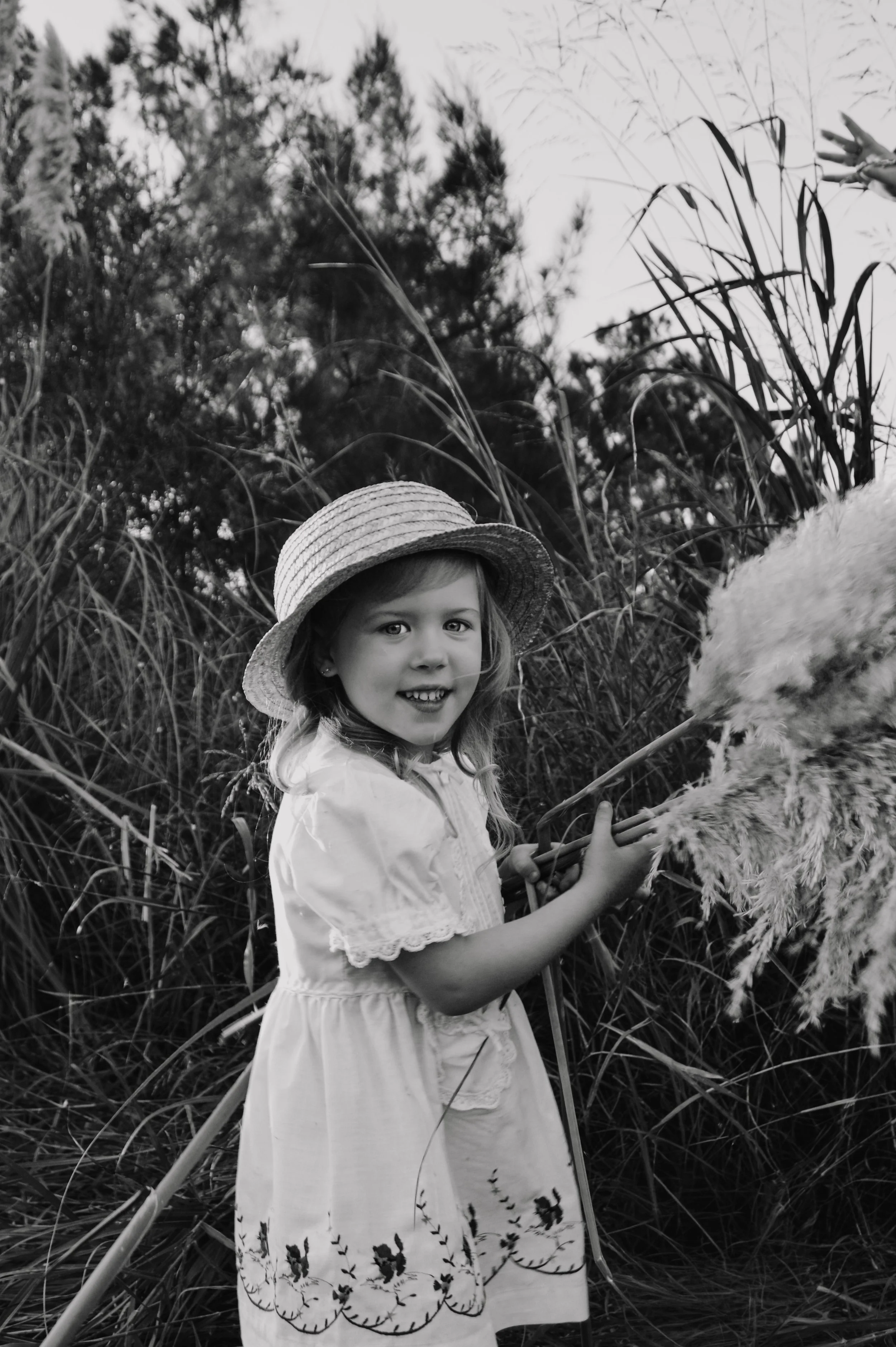 A young girl wearing a wide-brimmed straw hat and a dress with floral embroidery, standing outdoors among tall grass and trees, holding a fluffy flower with a smile.