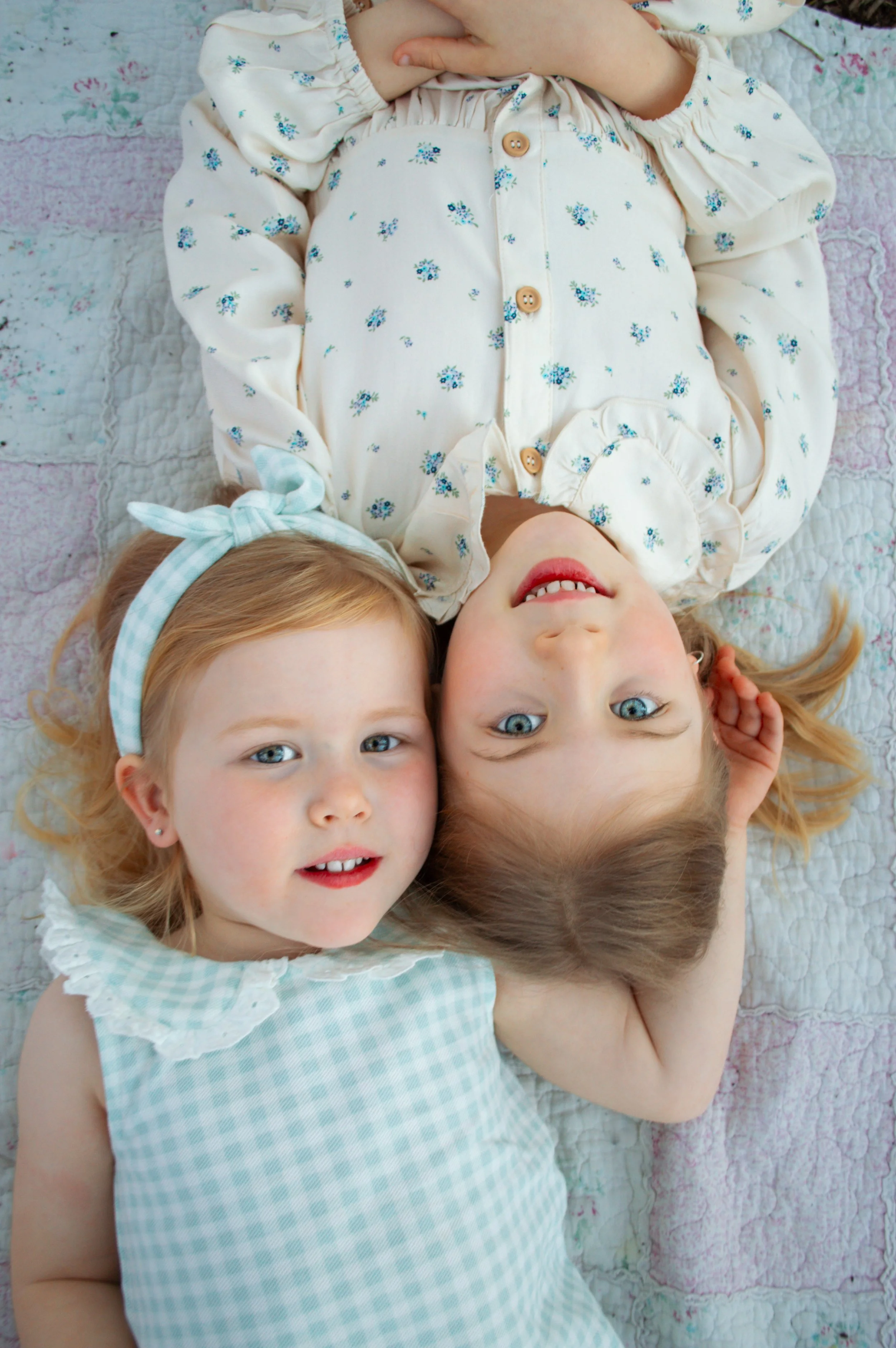 Two young girls lying on a quilted blanket, smiling at the camera. One girl has blonde hair with a light blue headband; the other girl has light brown hair.