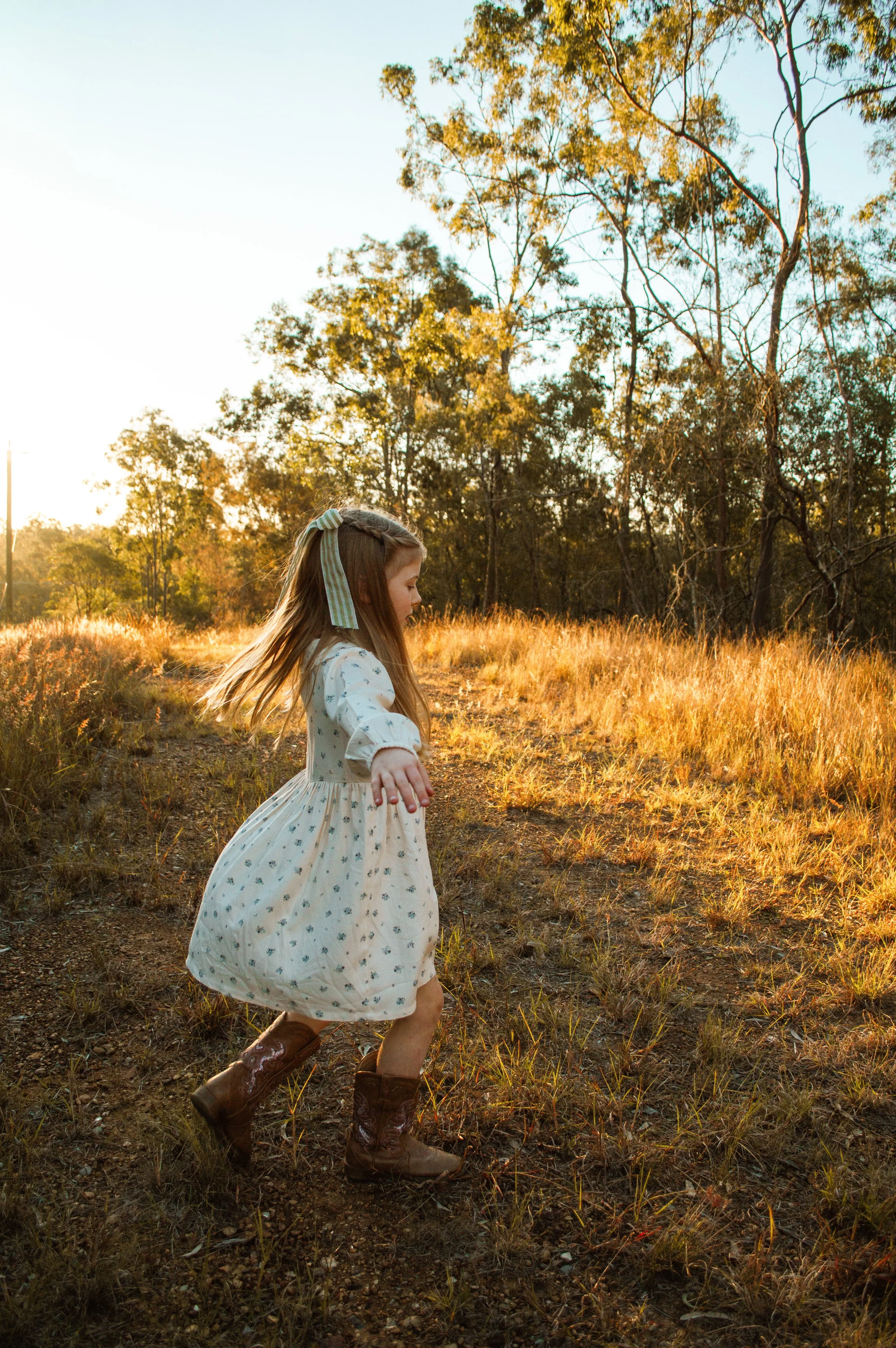 A young girl with long hair wearing a white dress with a floral pattern, brown boots, and a striped bow in her hair, running outdoors during sunset in a grassy field with trees in the background.