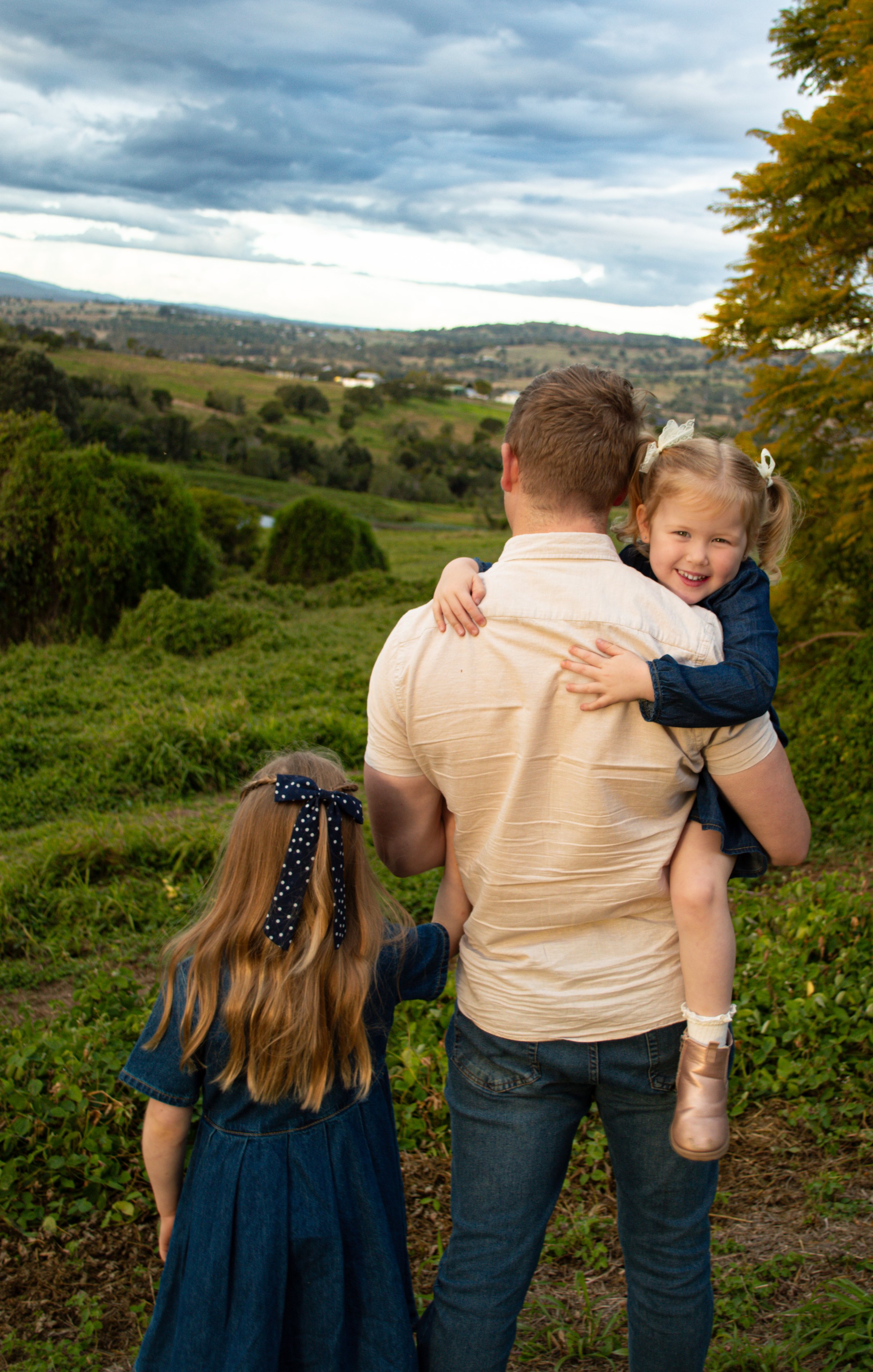 A man carrying a young girl on his back hugs her with a smile, while a young girl with long hair and a bow in her hair stands nearby. They are outdoors in a green landscape with trees, rolling hills, and a cloudy sky.