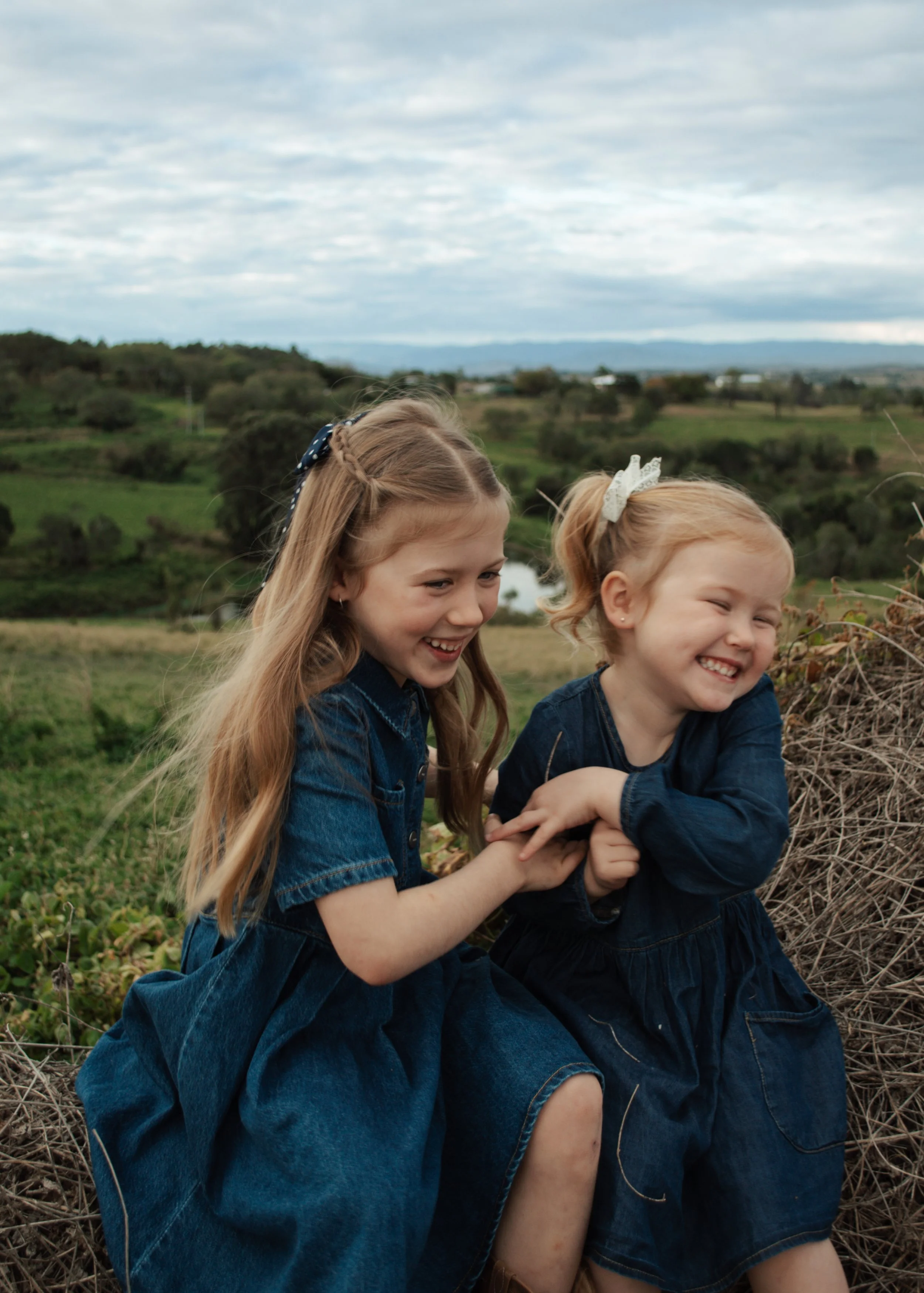 Two young girls sitting outdoors in a field, smiling and playing, with a scenic landscape of greenery, hills, and a cloudy sky in the background.