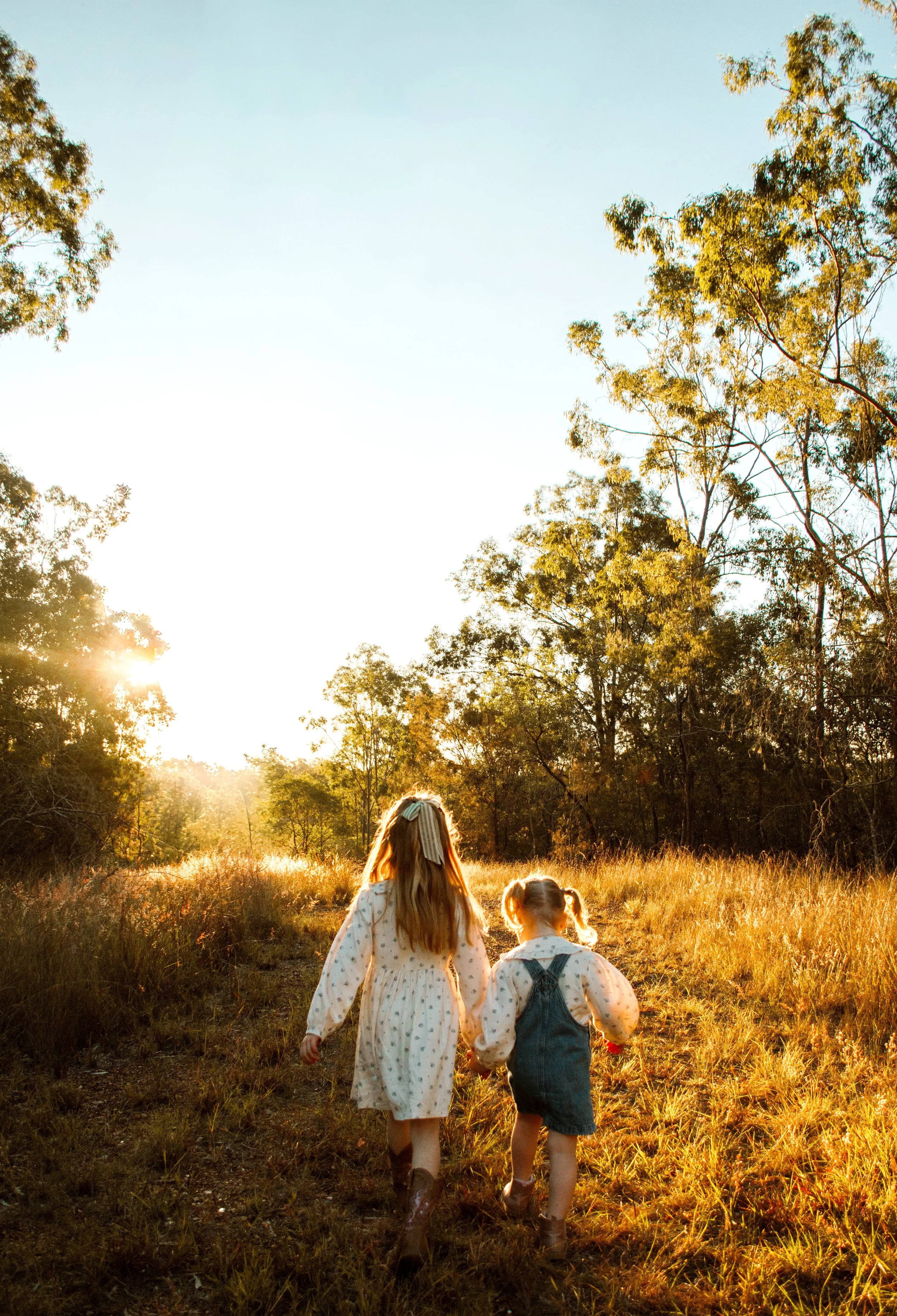 Two girls with long hair, wearing dresses and walking in a field during sunset or sunrise, with trees in the background.