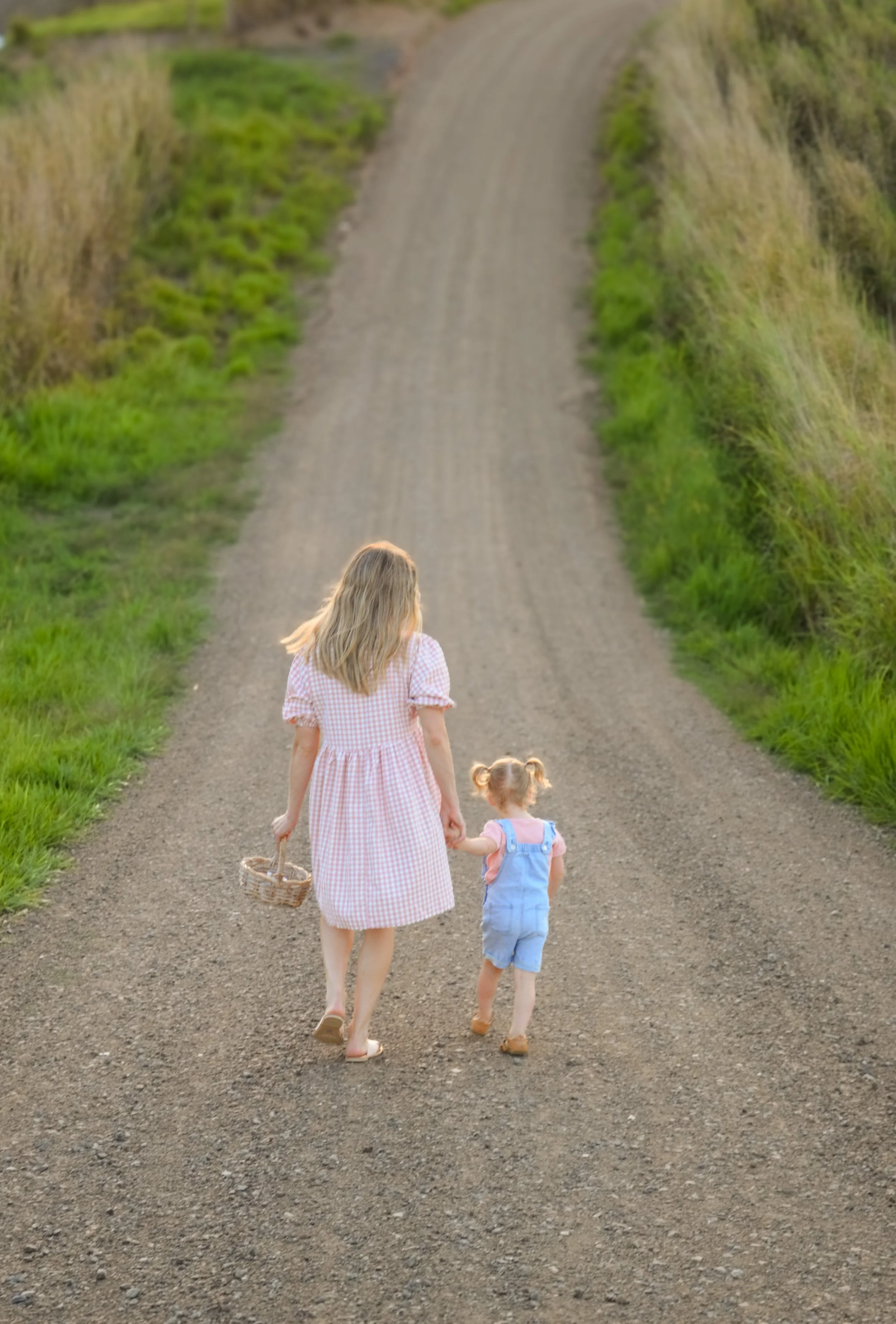 A woman and a young girl walk hand in hand down a dirt rural road surrounded by green grass and bushes, the woman holding a small basket.
