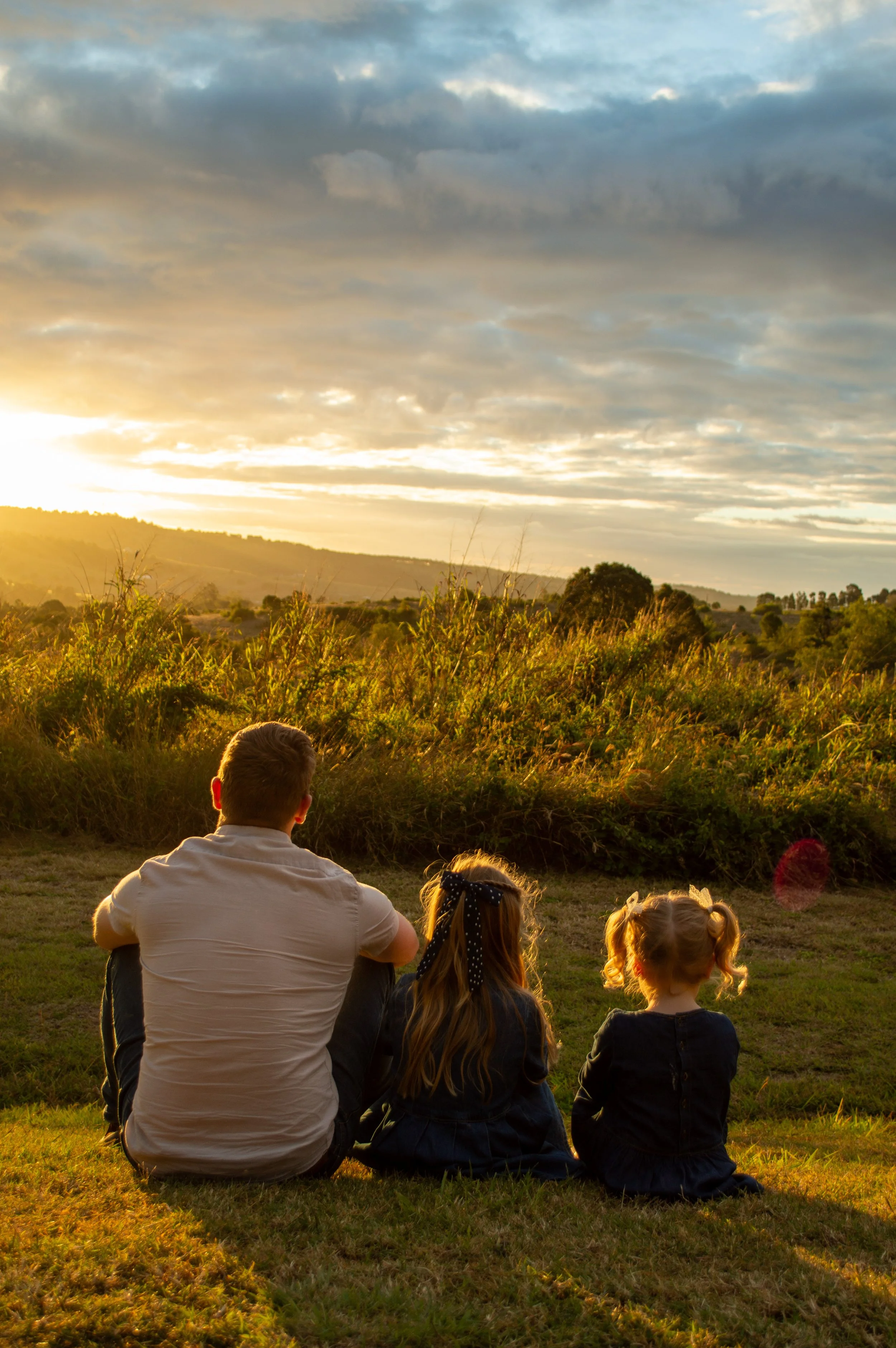 People sitting on grass and watching a sunset in a rural landscape with a cloudy sky.