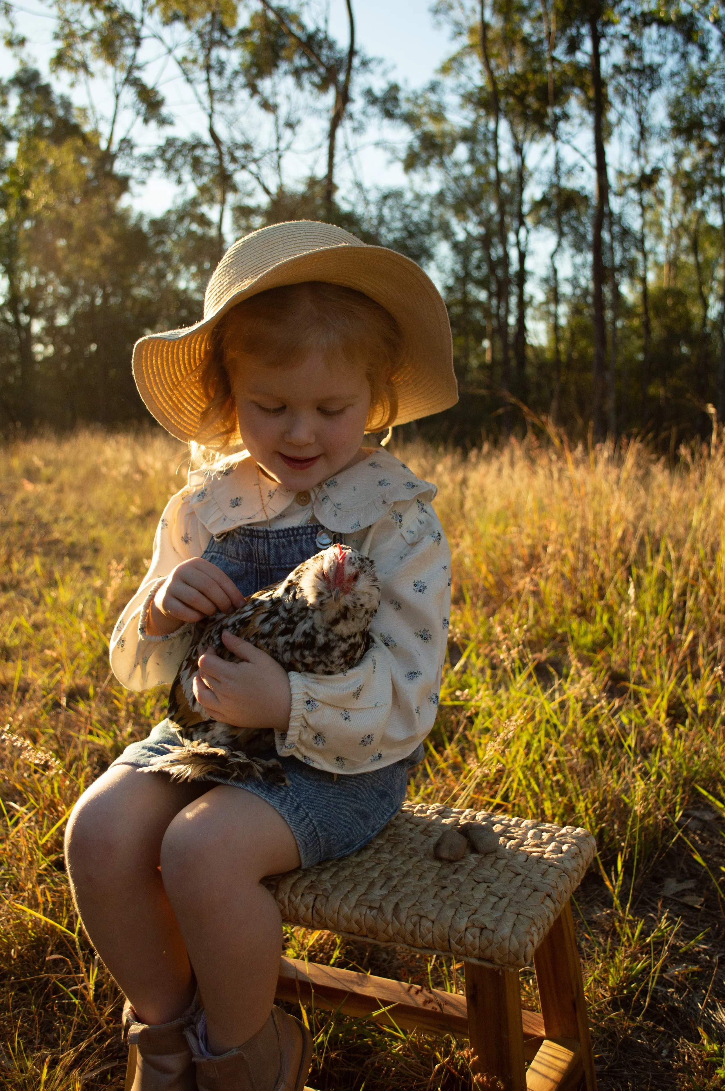 A young girl sitting on a woven stool outdoors in a grassy field, holding a small chicken. She wears a wide-brimmed hat, a long-sleeved shirt with floral details, denim shorts, and boots. The sunlight filters through trees in the background.