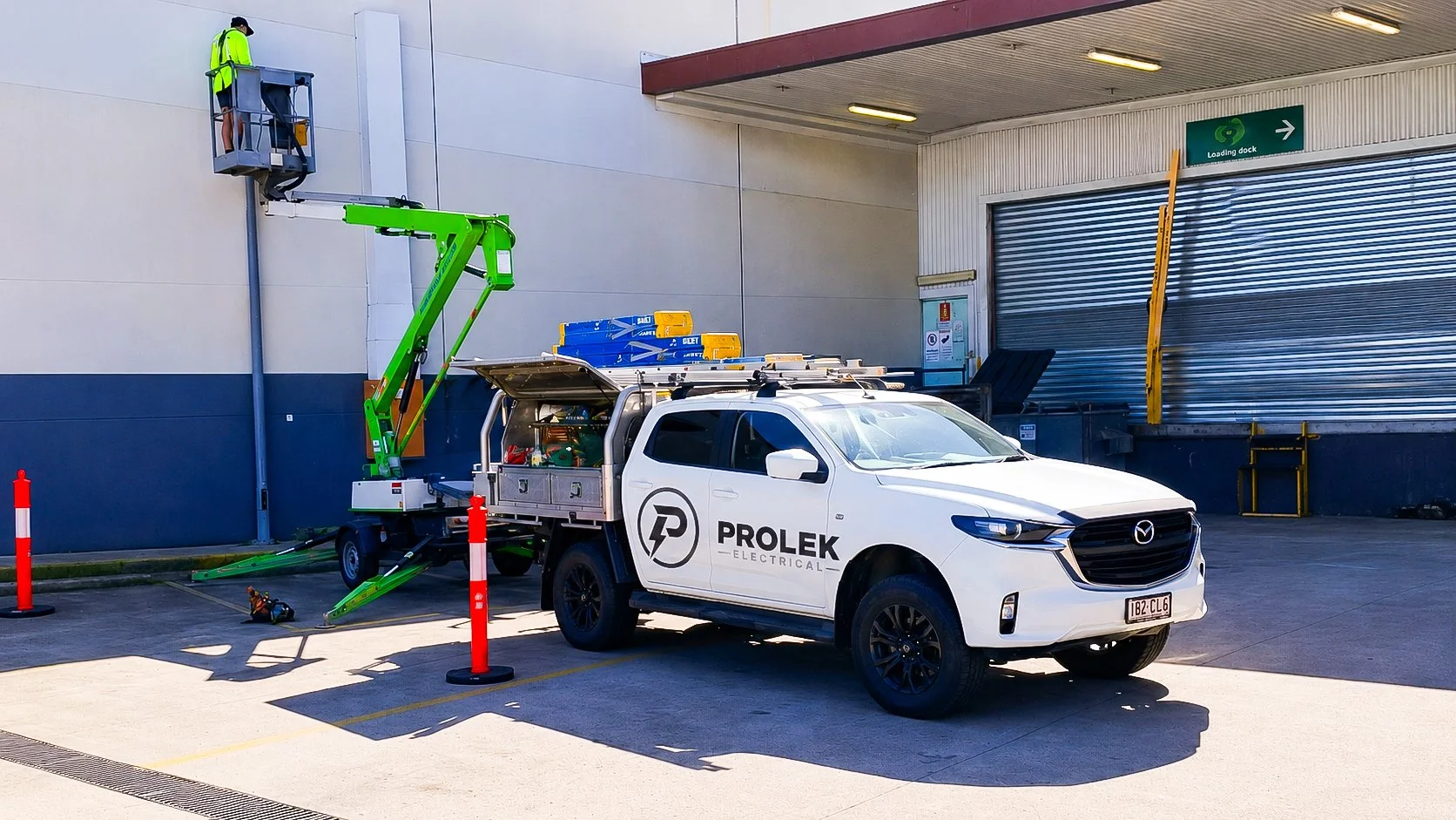 A white Prolek Electrical truck parked outside a loading dock, with a worker on a green lift working on the wall.