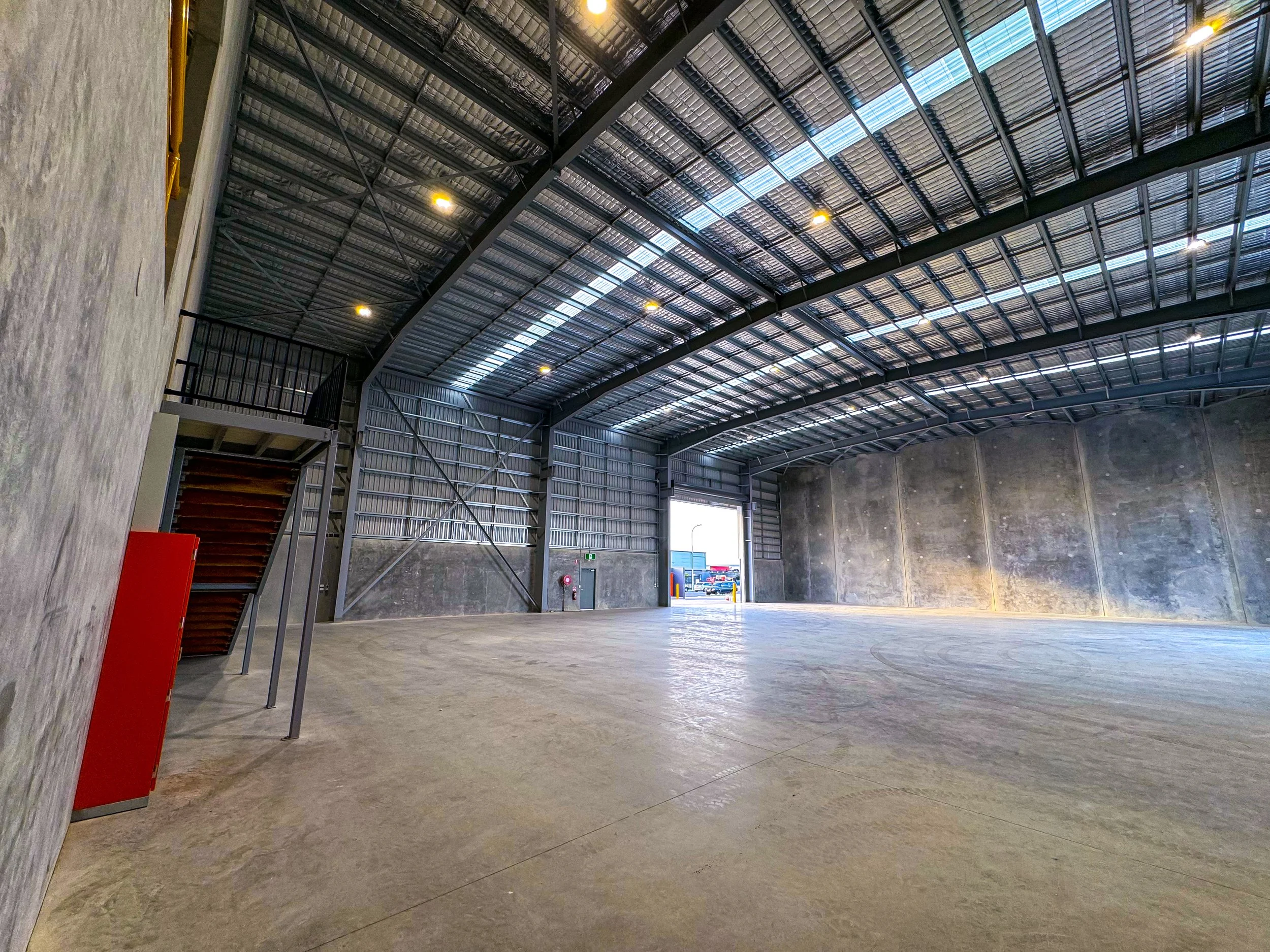 Interior view of a large empty industrial or warehouse space with high metal ceiling, concrete floor, and a open rolling door providing natural light and outside view.