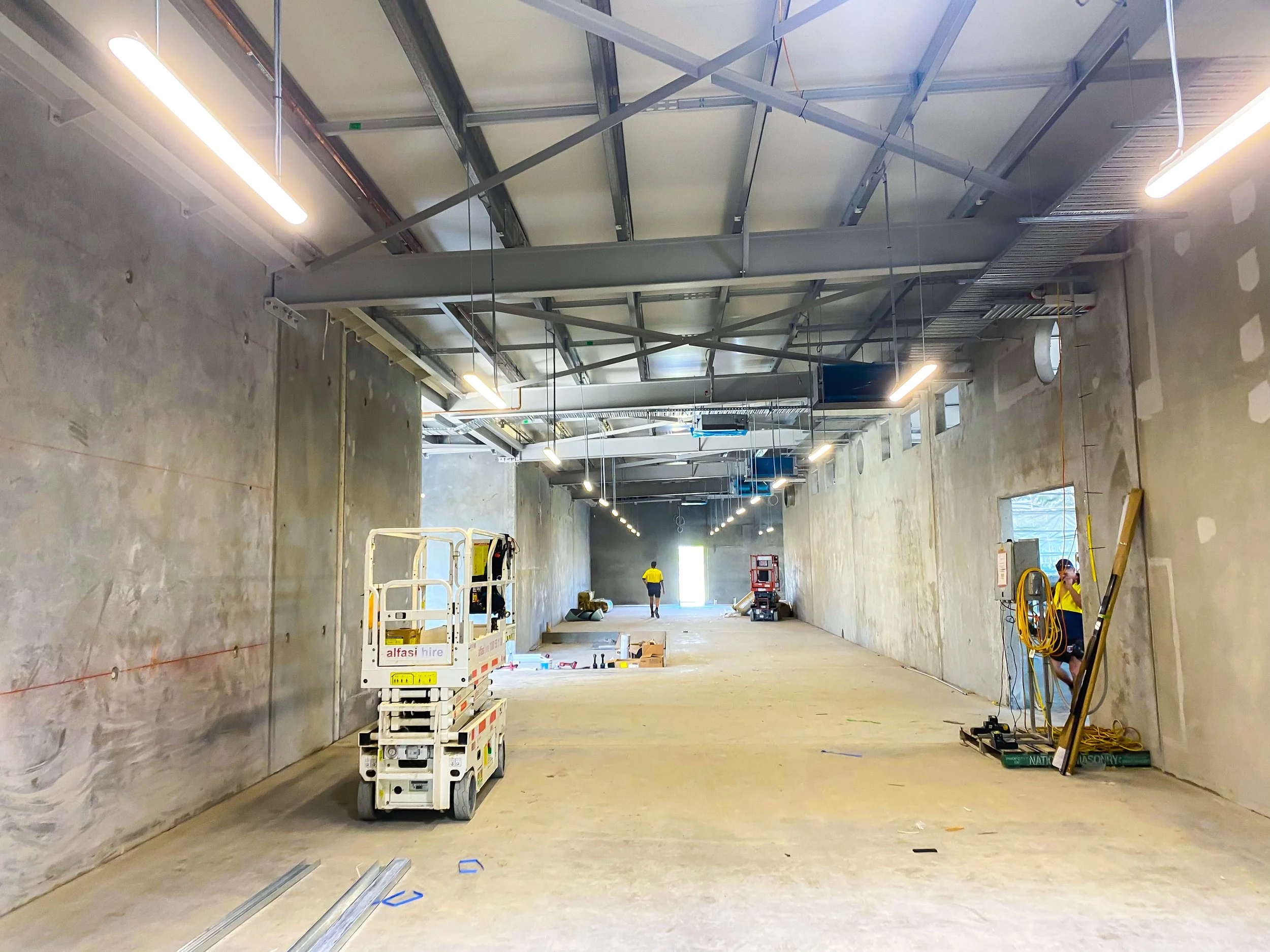 Construction site inside a large building with unfinished walls and ceiling. Workers are present, and construction materials and equipment are scattered throughout the space. Overhead lighting is installed, and the ceiling structure is exposed.