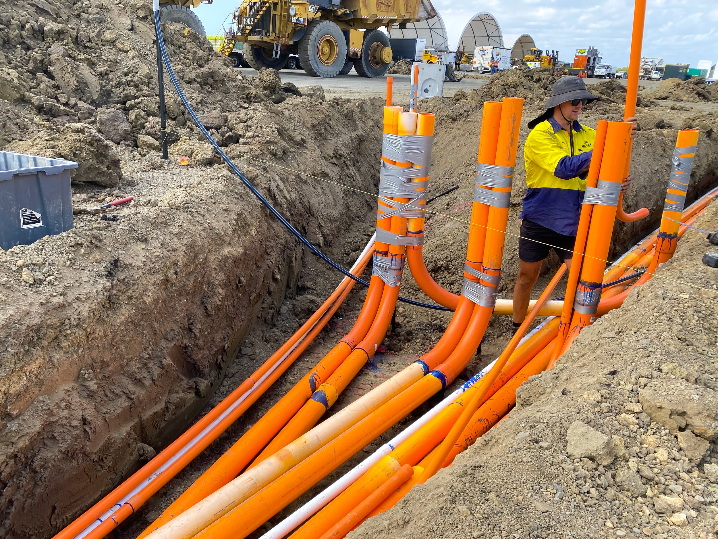 A construction worker wearing a yellow and blue jacket, a large wide-brimmed hat, and sunglasses stands among orange underground utility pipes in a deep trench at a construction site. The pipes are wrapped with gray tape, and various construction machinery and trucks are visible in the background under a cloudy sky.