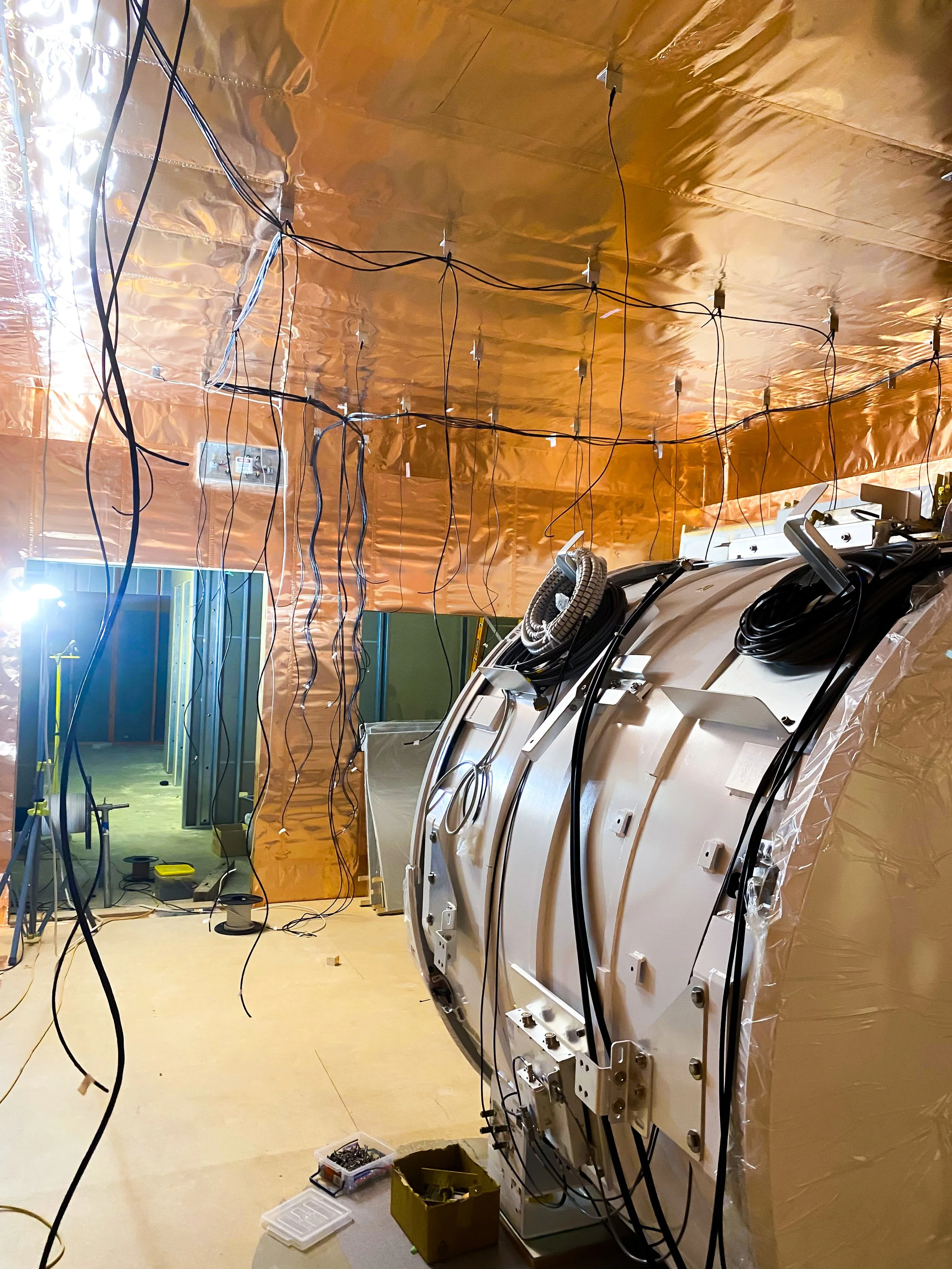 Construction site with a large rounded white structure to the right, covered in silver insulation material, surrounded by various black cables and electrical components. The background shows a room with green and yellow wall panels and a doorway with open framing.