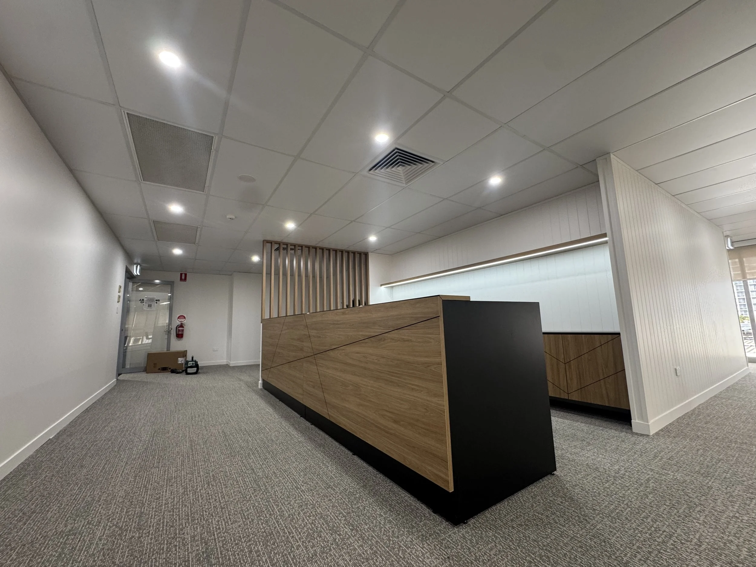Interior view of an office or lobby area with a reception desk, wooden panel decor, white walls, ceiling lights, and carpeted floor.