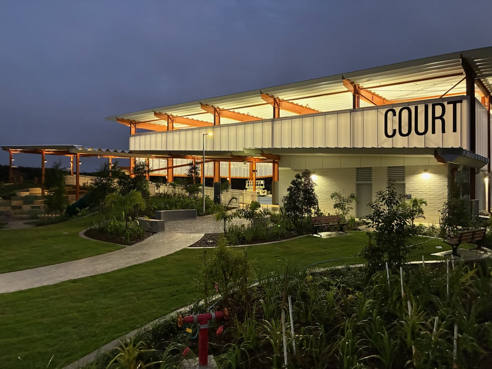 A modern courthouse building illuminated at night, featuring large glass windows, a prominent sign that reads 'COURT', landscaped garden with pathways, benches, and outdoor lighting.