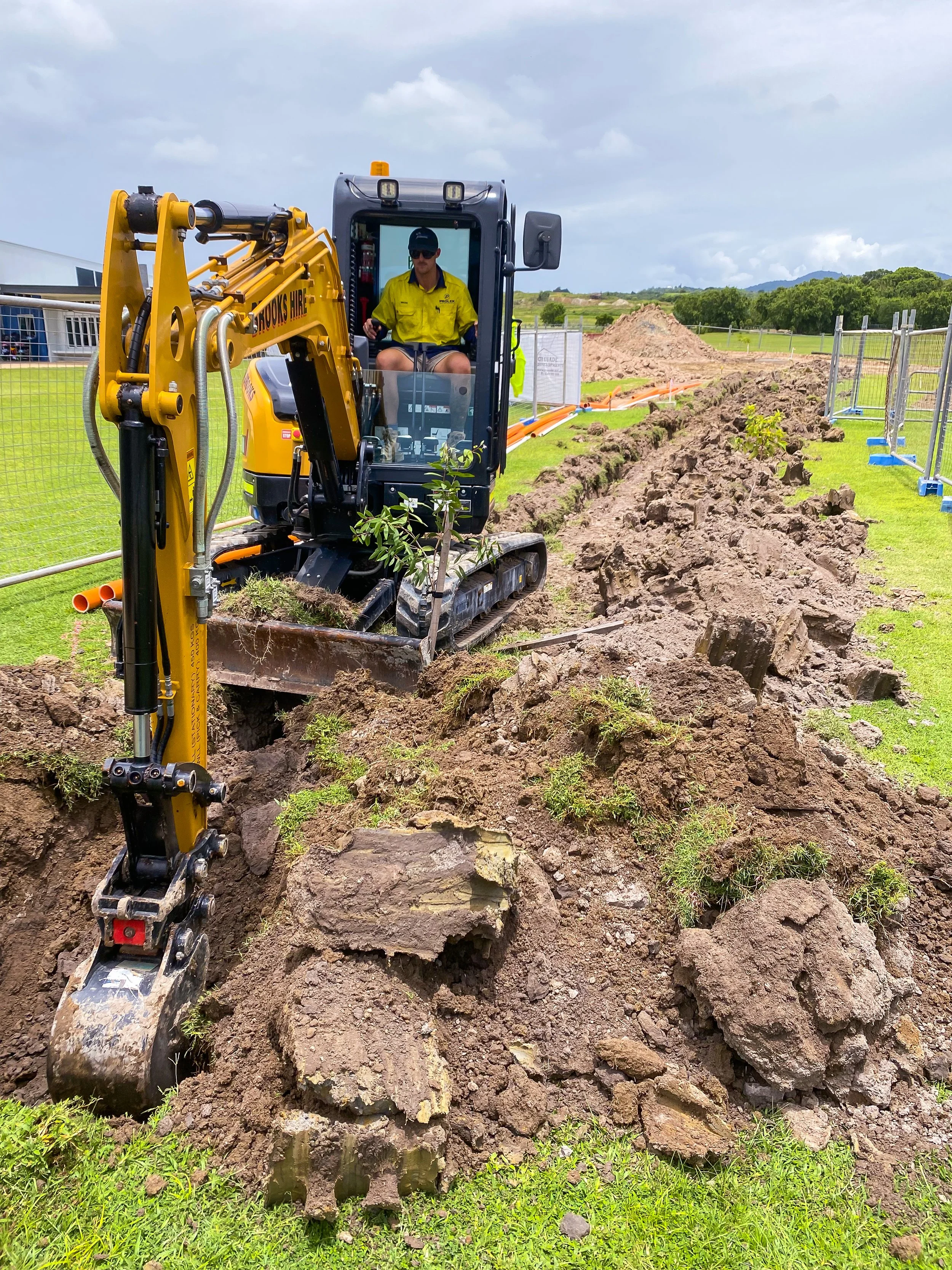 A person operating a small excavator digging a trench in a grassy field, with piles of dirt along the trench, surrounded by a metal fence and open landscape in the background.