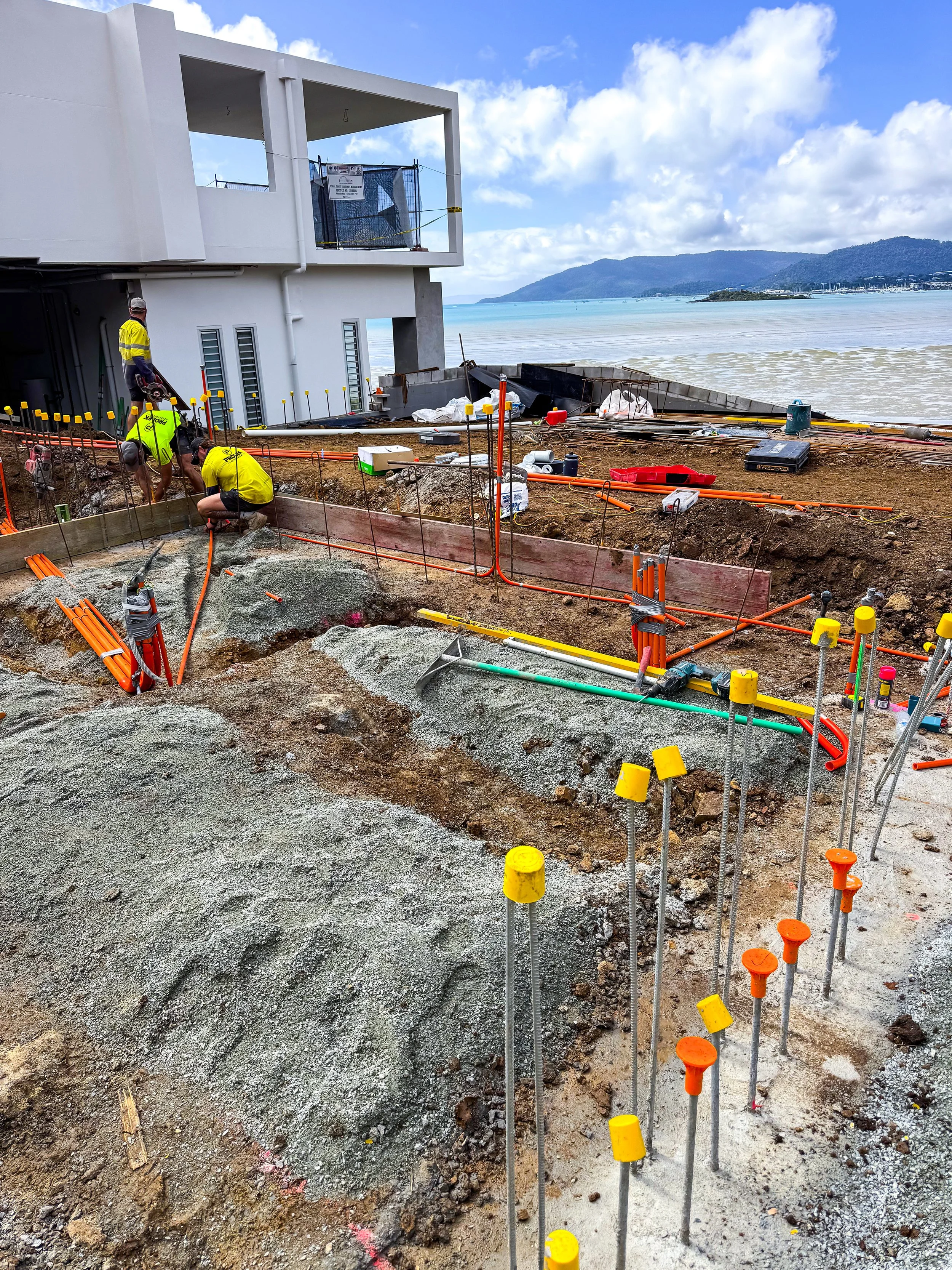 Construction workers working on a building foundation near the beach, with plumbing pipes and construction materials scattered around.