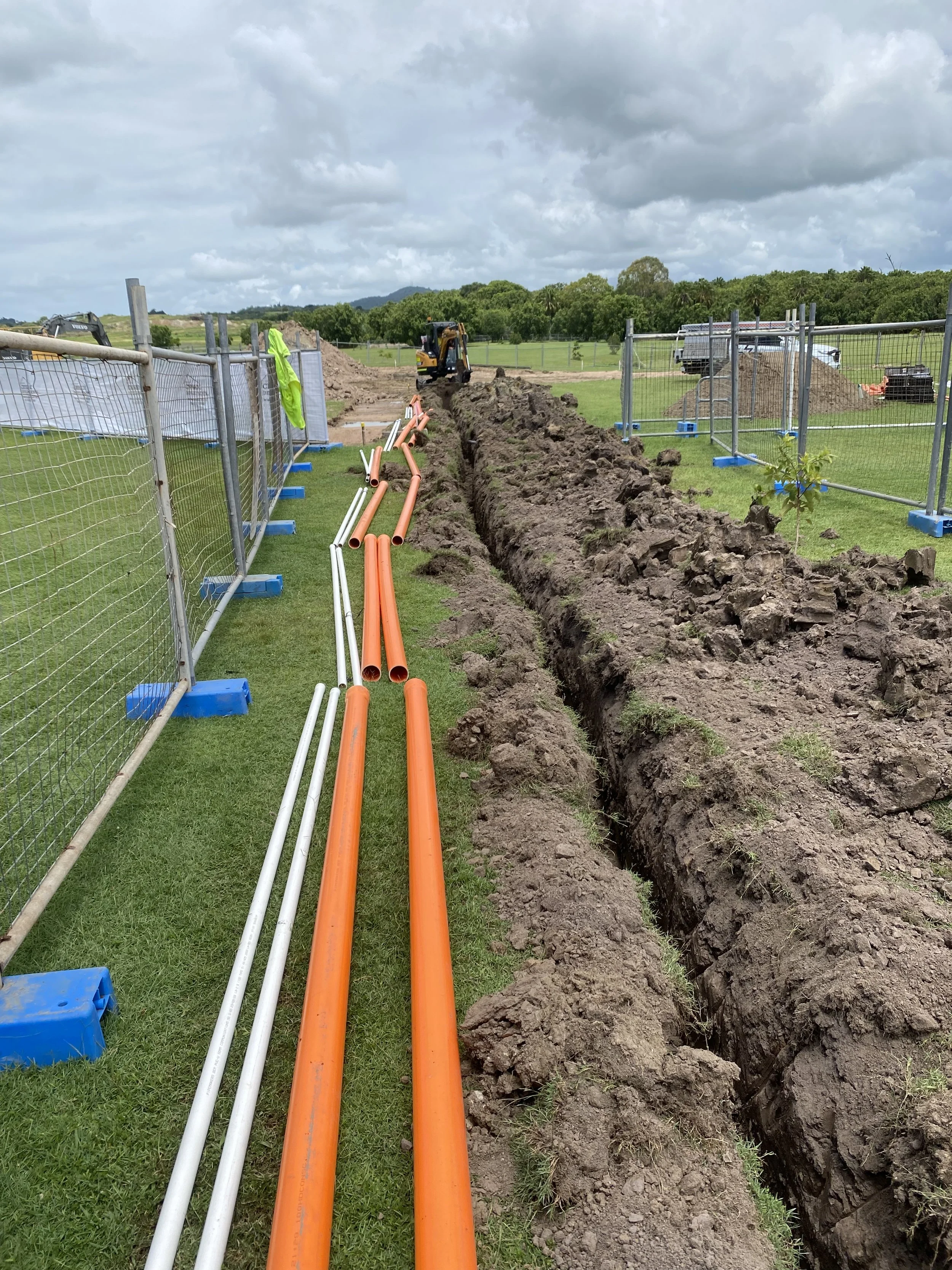 Construction workers are installing underground pipes in an open trench surrounded by a safety fence with grassy fields and cloudy sky in the background.