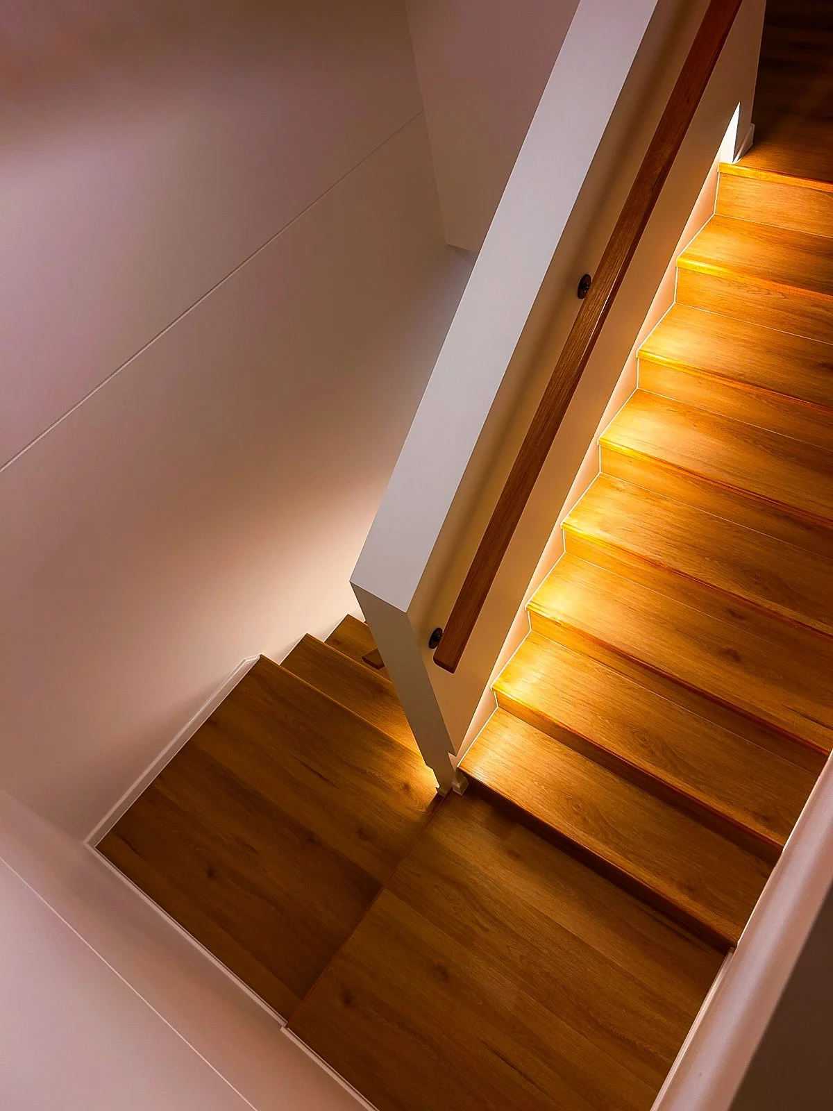 View of a staircase from above with wooden steps and built-in lighting along the edge, railings on the side, and a beige wall.
