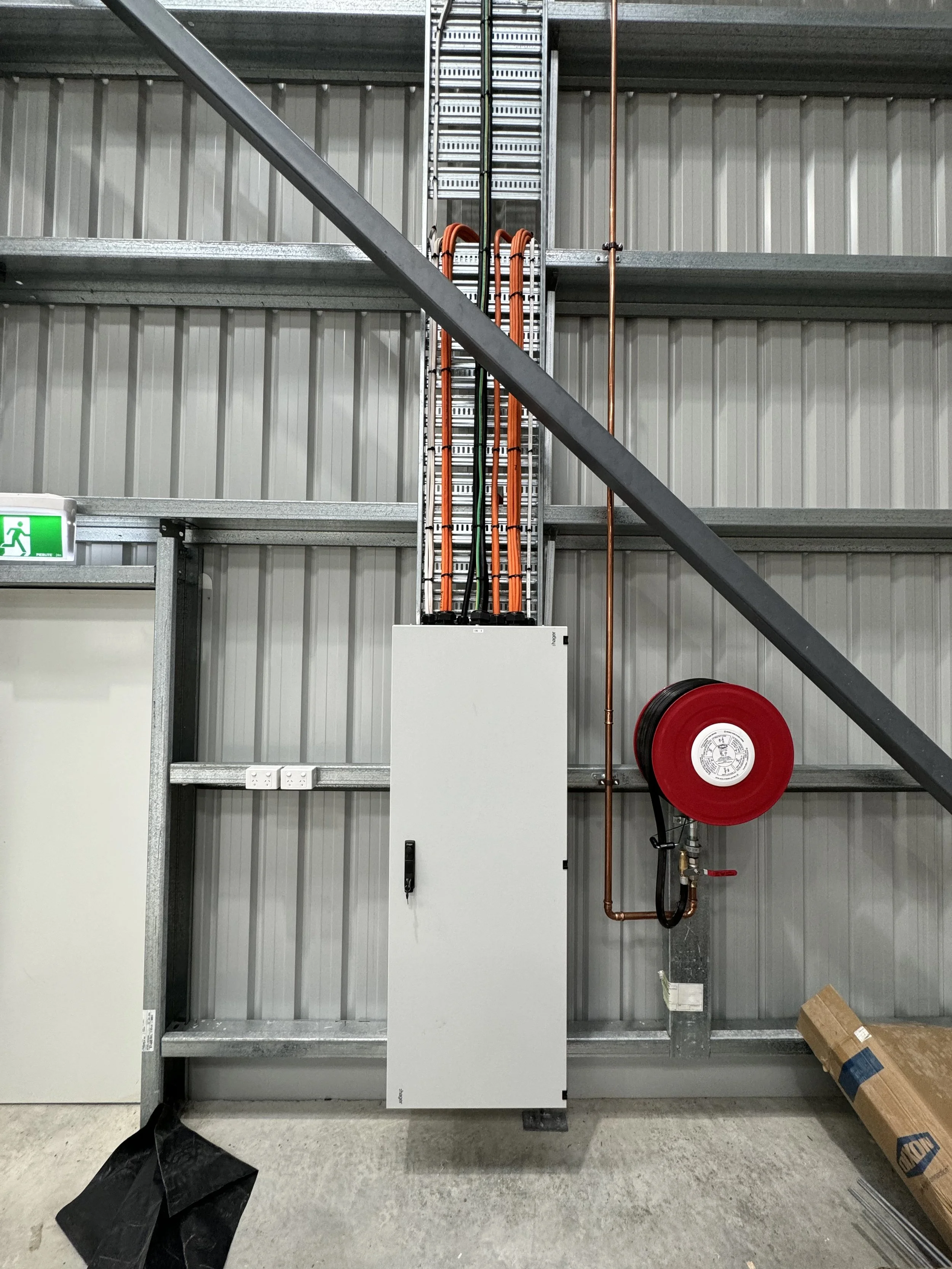 The image shows a warehouse with a metal wall and ceiling. There is a wire shelving unit, orange cables, a copper pipe, a fire hose reel, electrical outlets, and some tools and boxes on the ground.