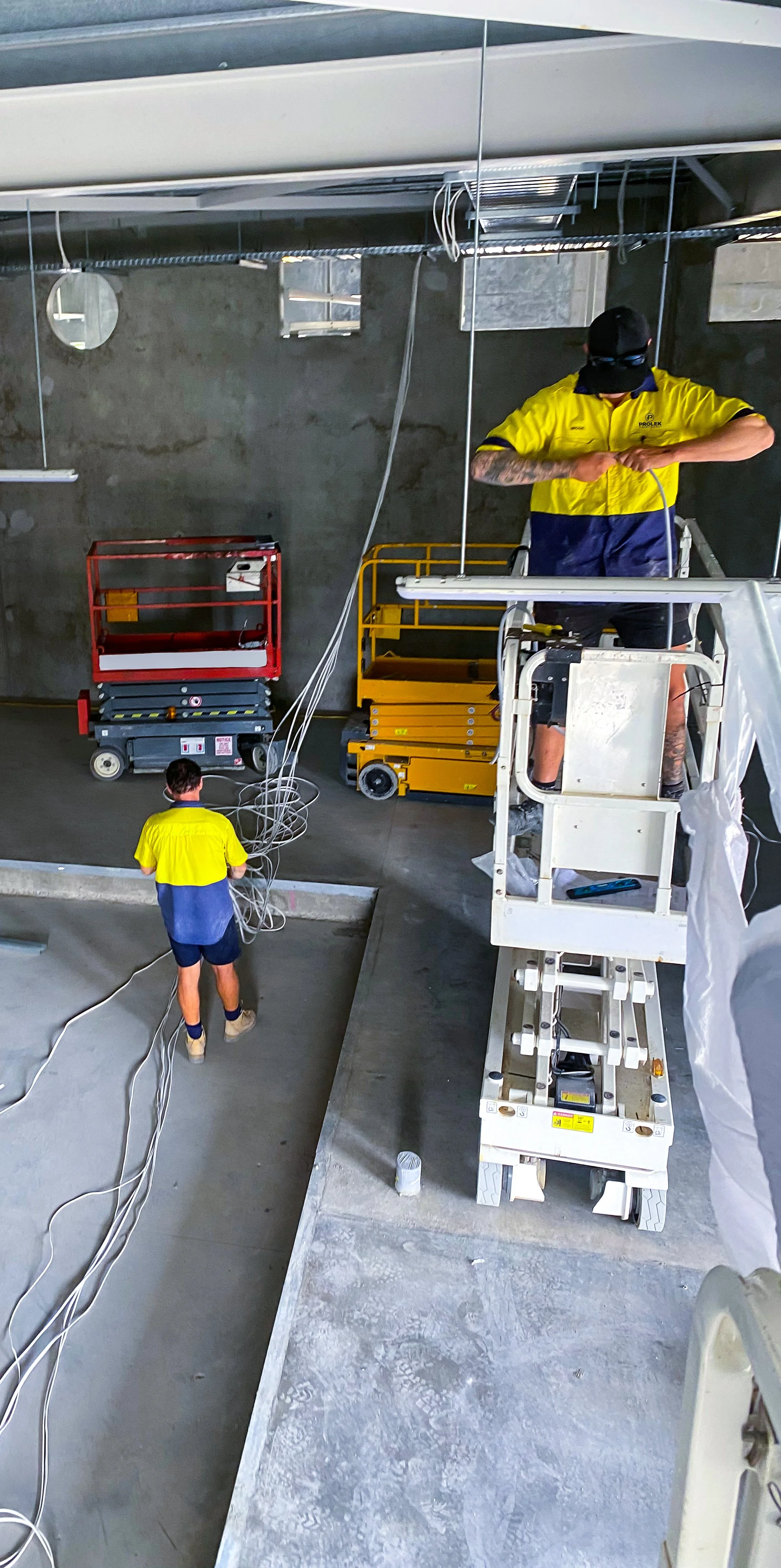 Two workers in yellow shirts are working on electrical wiring in a construction site. One is on a scissor lift, and the other is on the ground holding cables. There are various construction tools and equipment around, with a concrete wall in the background.