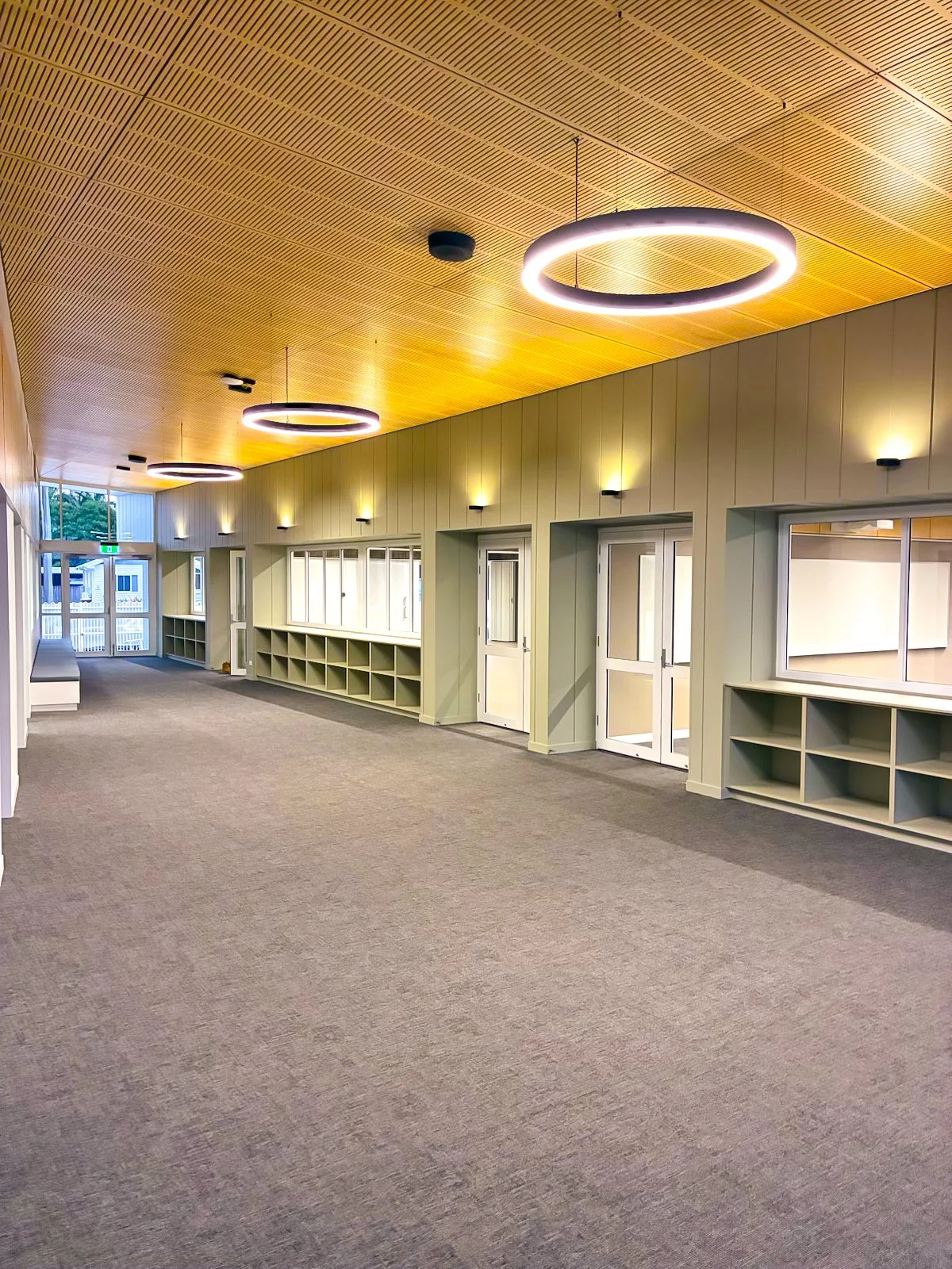 Bright interior hallway with wooden ceiling, circular ceiling lights, and built-in shelves along the walls. The hallway leads to a door with glass panels and has large windows letting in natural light.