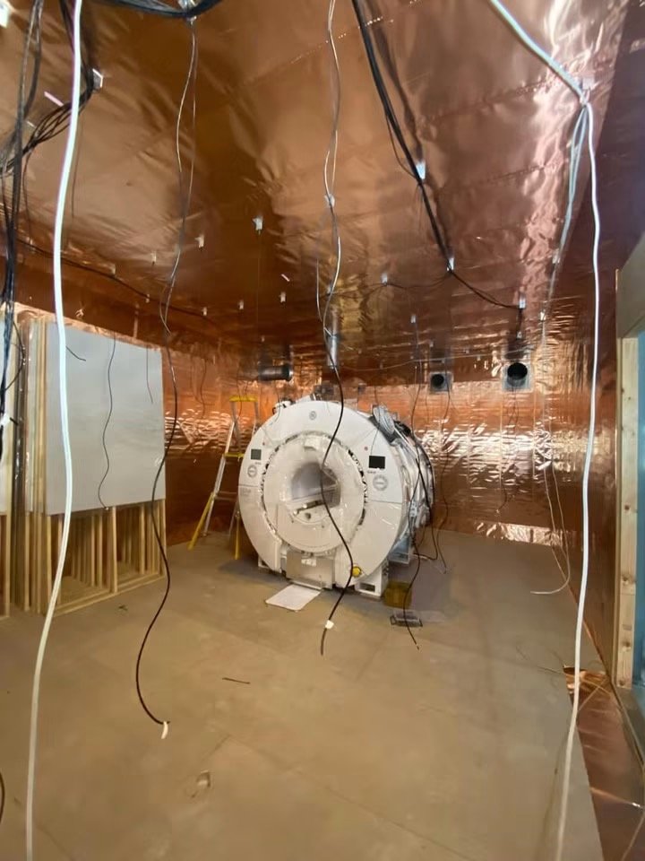 A blank room with an MRI machine, copper-colored wall coverings on the ceiling and walls, and loose wires hanging from the ceiling.