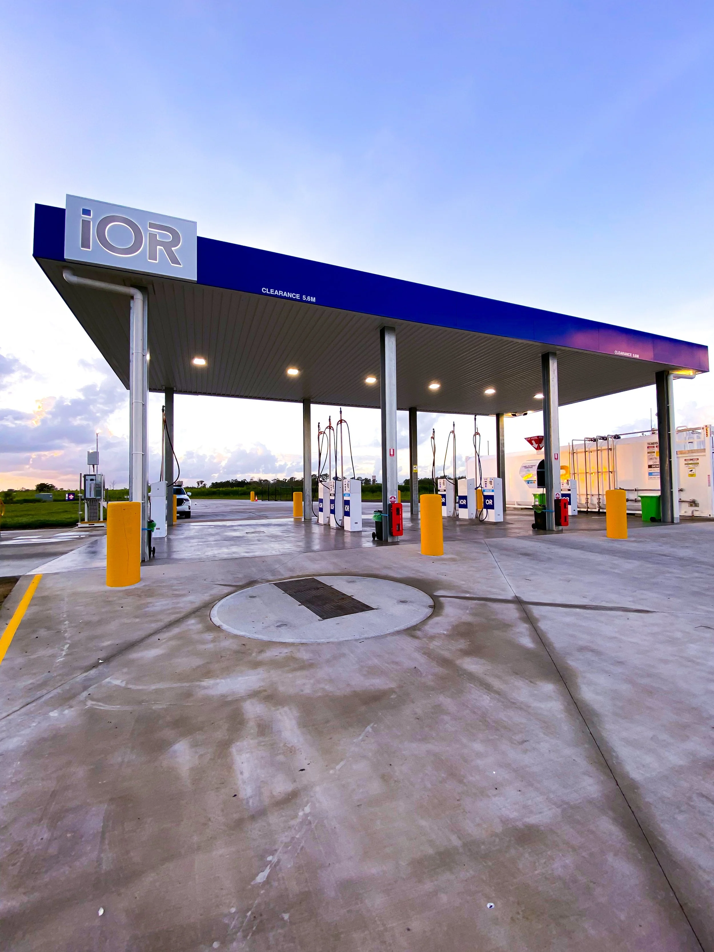 Electric vehicle charging station with multiple chargers under a blue canopy, surrounded by a parking lot and open landscape during sunset.