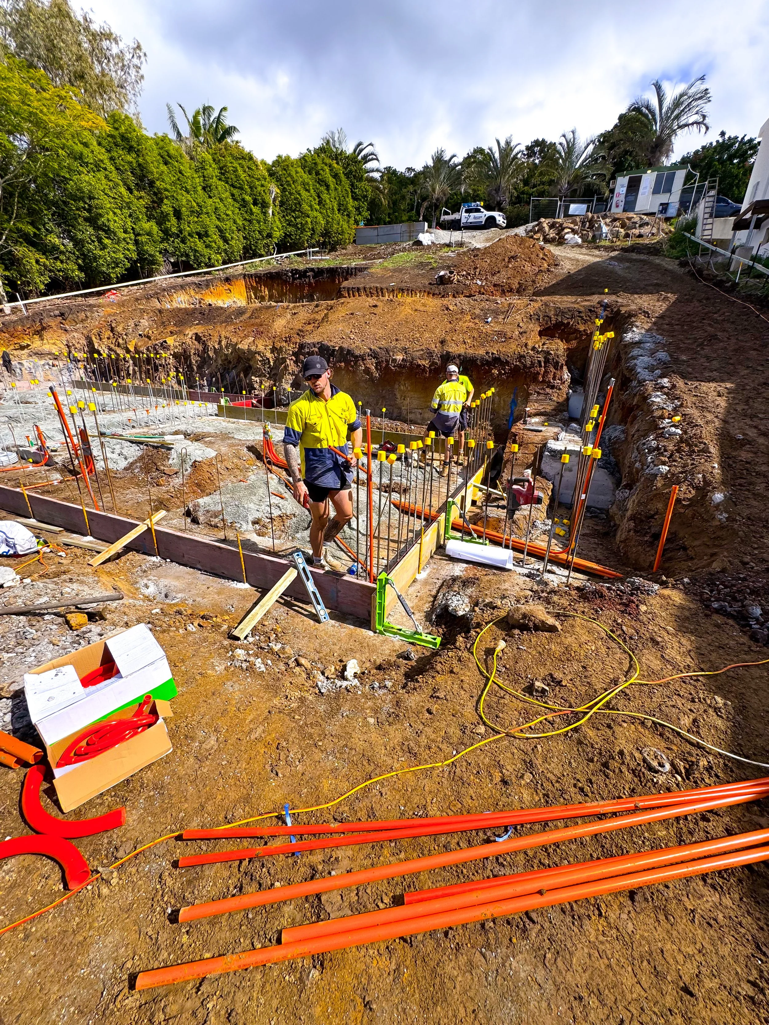 Construction workers on an excavation site installing underground piping. Metal rebar structures are in place, with orange pipes arranged on the ground, and tools and equipment scattered around.